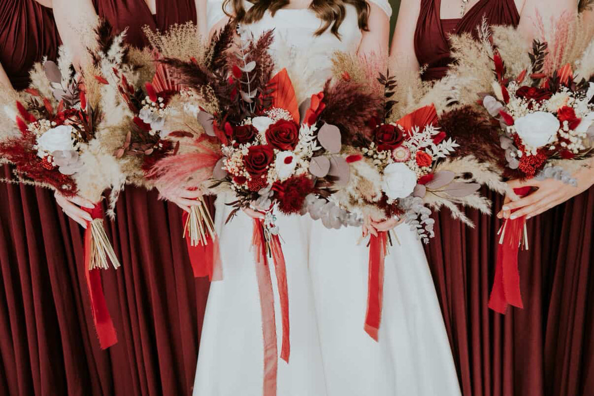 Bride holding a bouquet with red and white flowers, surrounded by bridesmaids in burgundy dresses.