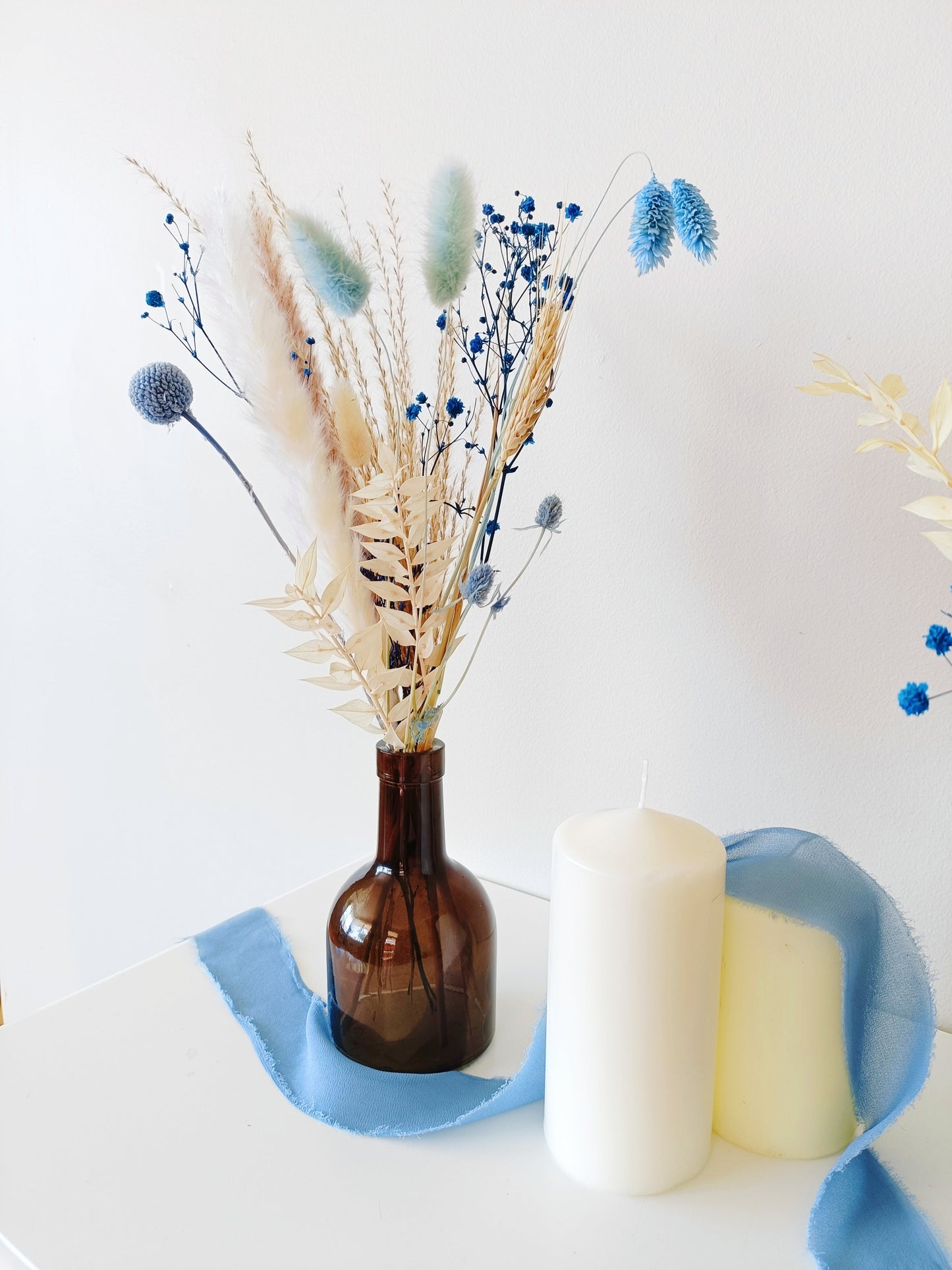 Dried flower arrangement in brown glass vase with blue and white accents – styled with candles for coastal home or event decor.