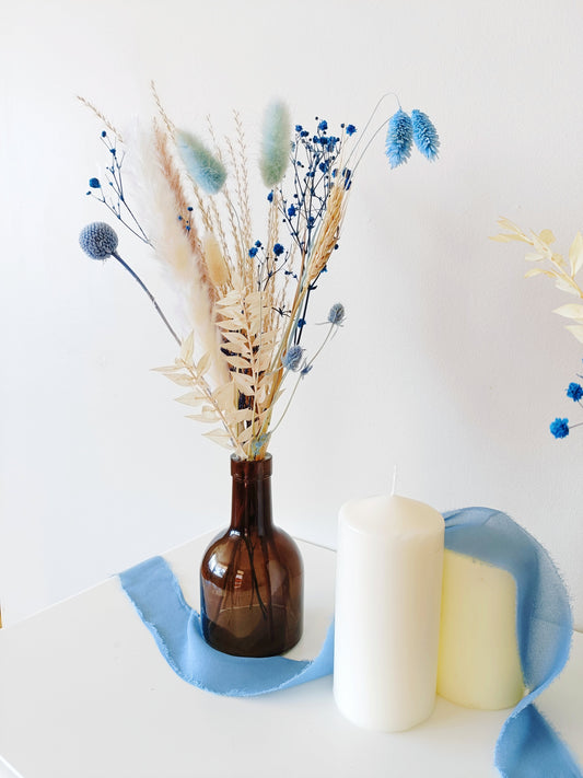 Dried flower arrangement in brown glass vase with blue and white accents – styled with candles for coastal home or event decor.