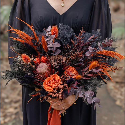 Bridal bouquet in burnt orange and black tones featuring preserved roses, Australian native flowers, textured foliage, and dried grasses, held by a woman in a black dress for a modern boho wedding.