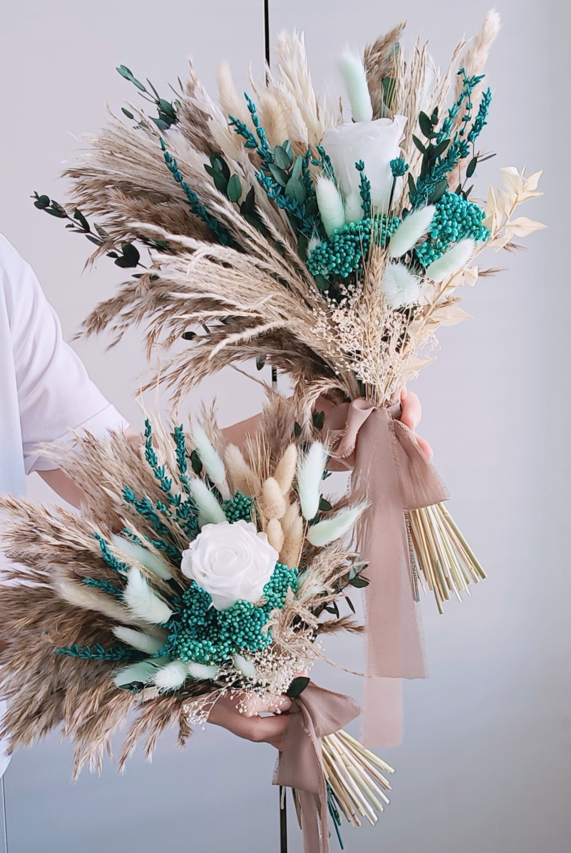 Featuring a sophisticated mix of teal and white, this dried flower bouquet is a striking choice for the modern boho bride. Pampas grass and preserved foliage add a touch of natural elegance, while the teal accents bring a fresh and vibrant feel. Perfect for rustic-themed weddings, this long-lasting bouquet is designed to make a lasting impression. Discover more teal and white bridal bouquets in our exclusive collection.