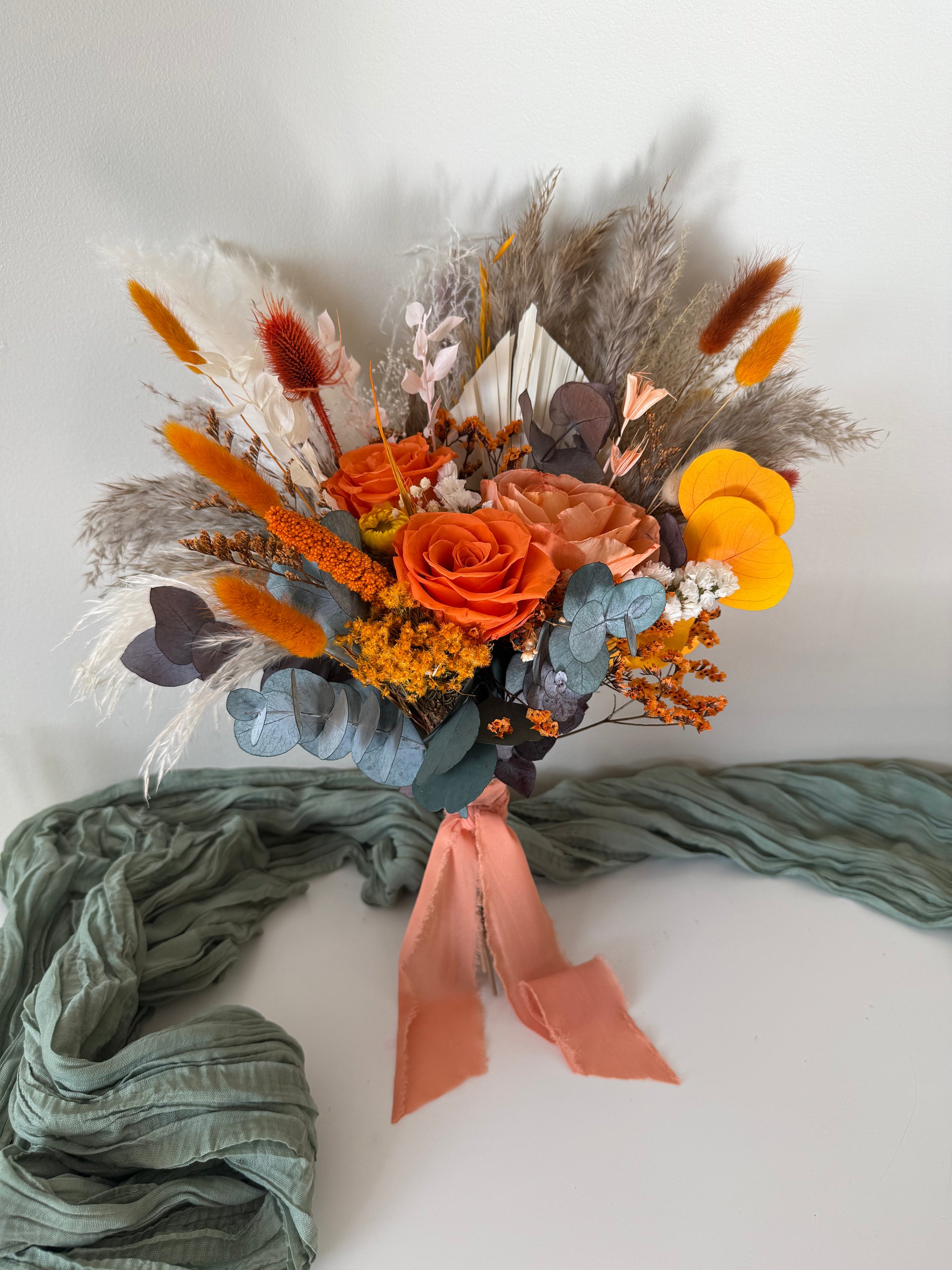 Boho bridal bouquet with terracotta, orange, and yellow dried flowers, sola wood roses, pampas grass, and eucalyptus, tied with a dusty pink ribbon, styled with green fabric.