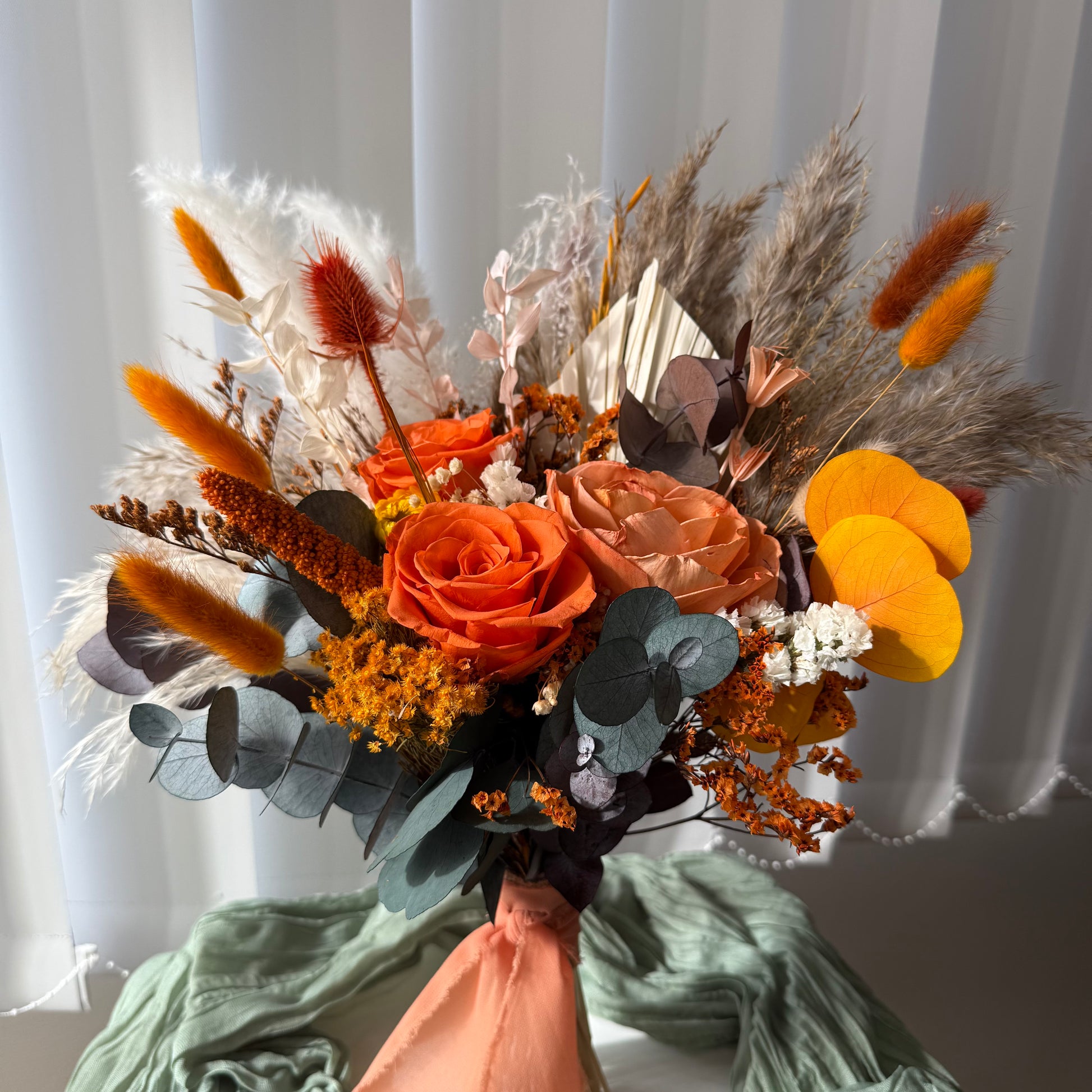 Side view of a preserved flower bouquet with terracotta roses, yellow accents, dried wheat, and bunny tails, tied with a soft pink ribbon for boho weddings.