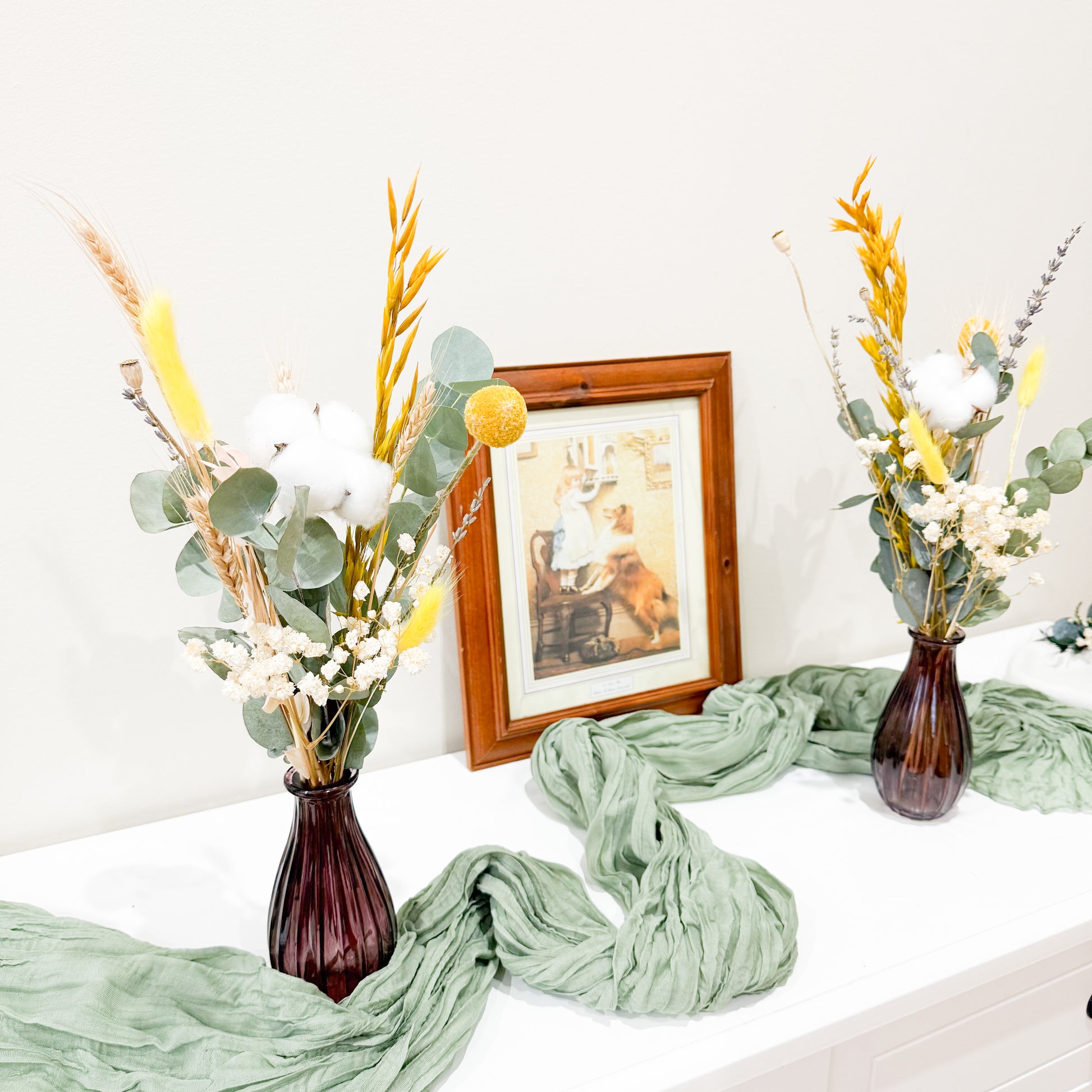 Decorative setup with vases of Australian flowers, green fabric, and a framed picture on a white surface.