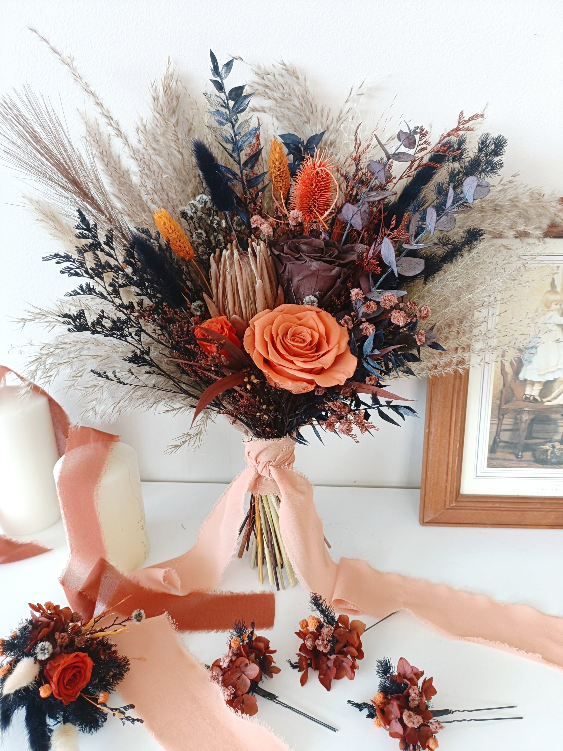 Dried flower bouquet in burnt orange and navy tones with rose centerpiece, paired with coordinating boutonniere and hair accessories on white background.