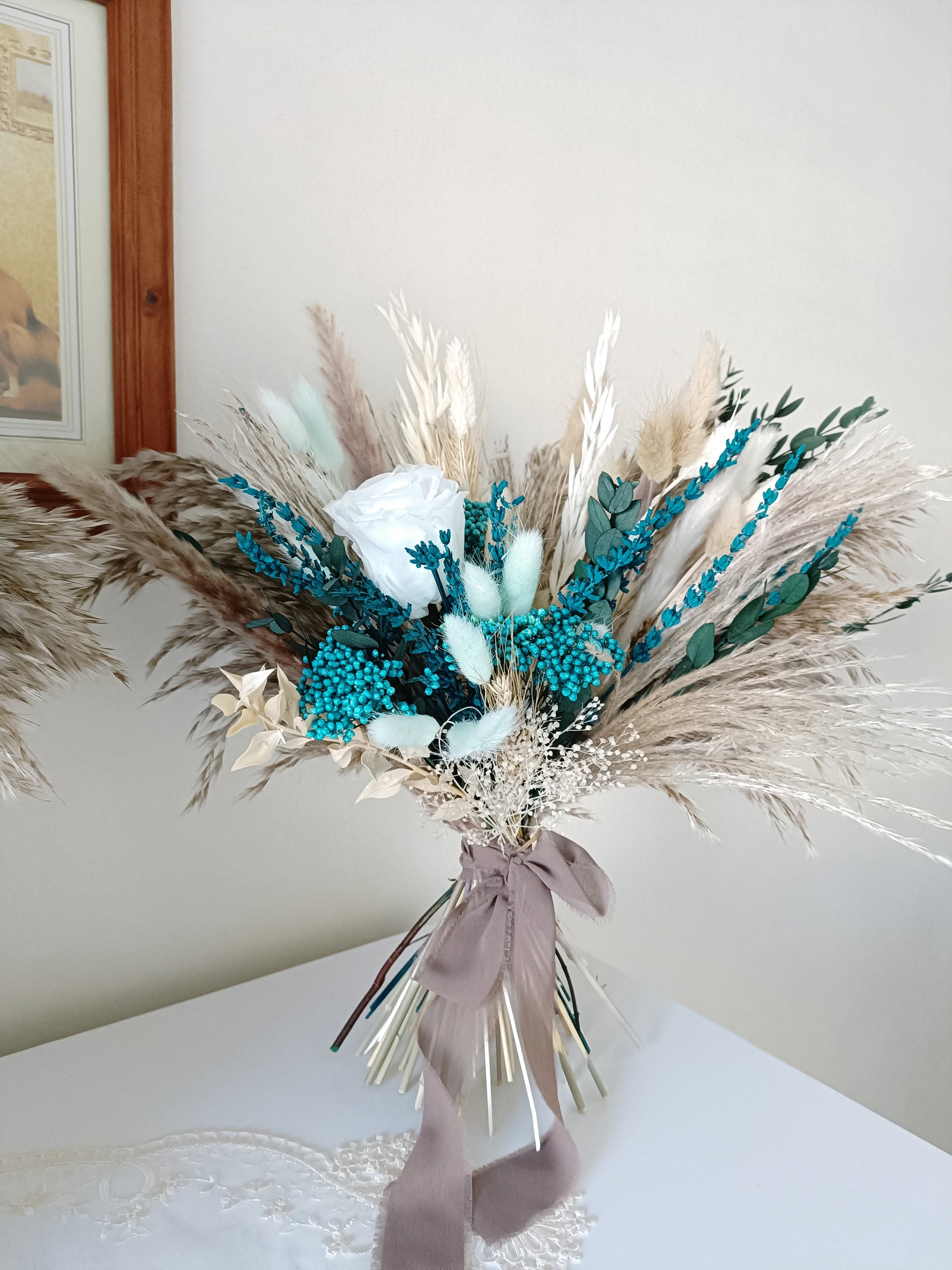 This beautifully crafted wedding bouquet features an elegant mix of teal, white, and natural tones, made from dried flowers and pampas grass. The arrangement exudes a modern rustic charm, with preserved foliage and delicate accents adding a sophisticated touch. Perfect for a contemporary bride, this long-lasting bouquet is ideal for weddings with a natural or bohemian theme. Explore our collection for more unique bridal bouquets and wedding decor inspirations.
