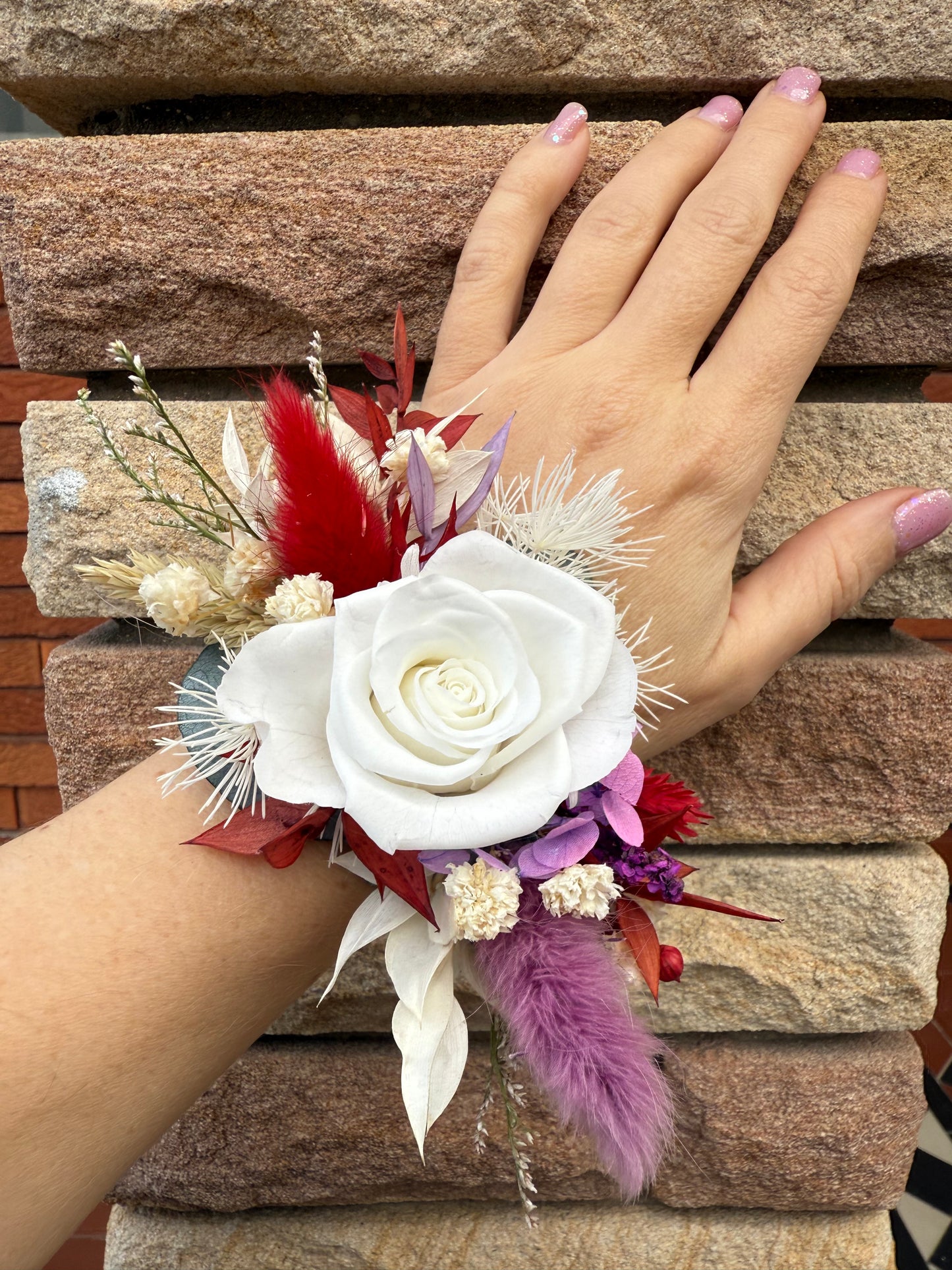 Hand wearing a wrist corsage with white flowers and purple and red accents on a wooden surface.