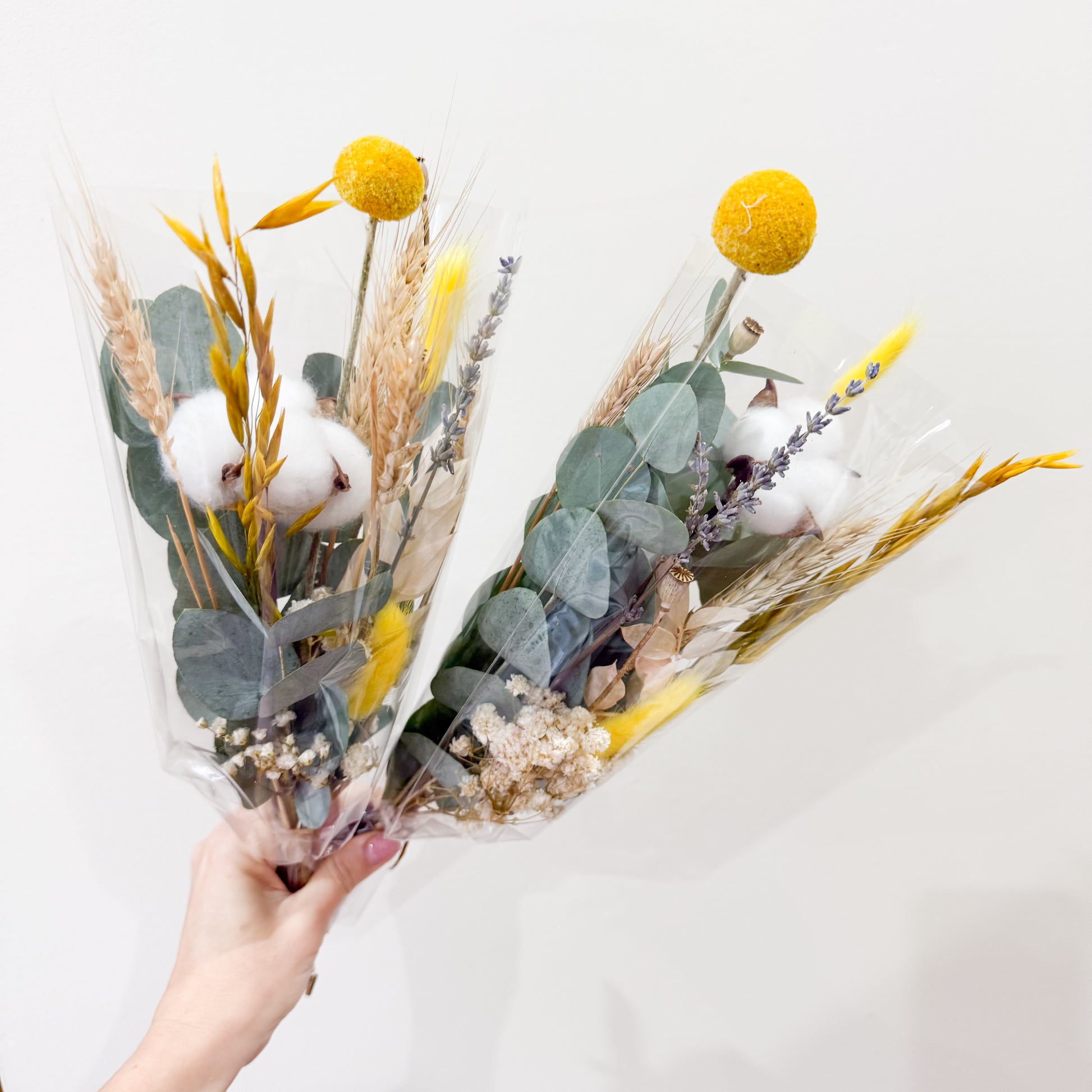 Handheld dried bouquet with preserved eucalyptus, yellow craspedia, cotton, wheat, and rustic grasses against a red brick wall.