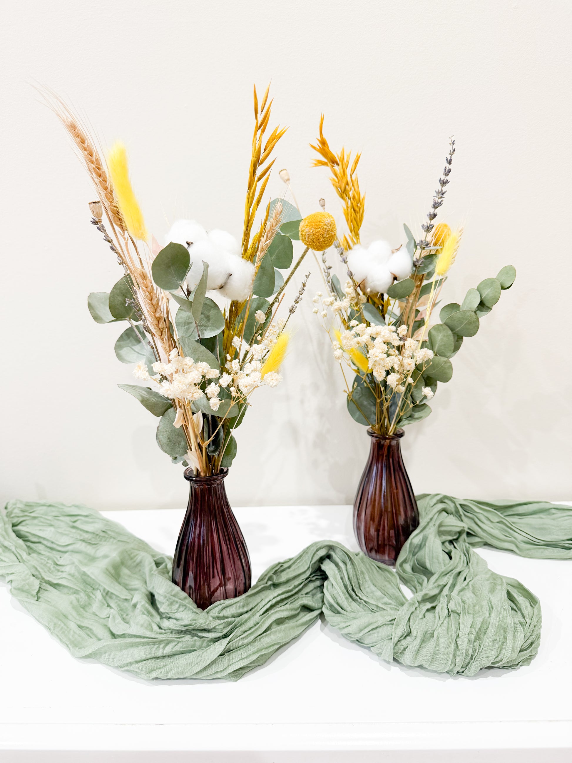 Pair of dried flower arrangements in brown vases styled with green fabric, including eucalyptus, bunny tails, and billy buttons.