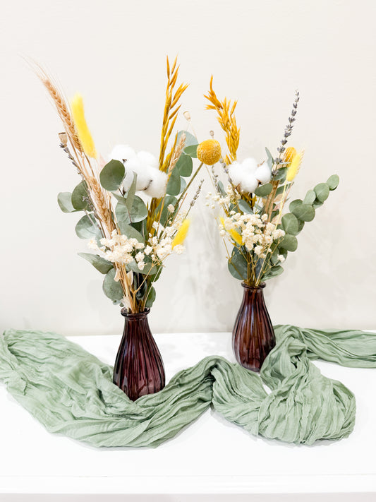 Pair of dried flower arrangements in brown vases styled with green fabric, including eucalyptus, bunny tails, and billy buttons.