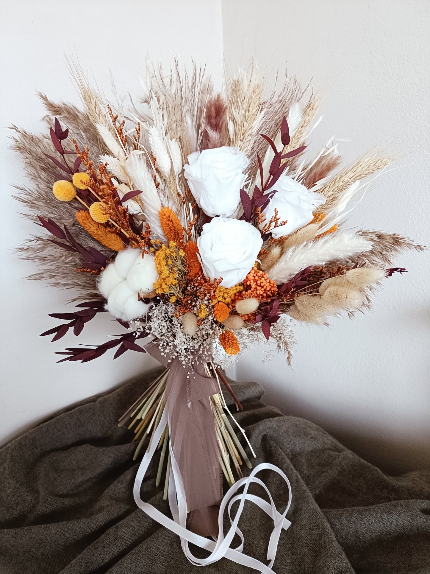 This beautifully crafted wedding bouquet features an elegant mix of orange, white, and natural tones, made from dried flowers and pampas grass. The arrangement exudes a modern rustic charm, with preserved foliage and delicate accents adding a sophisticated touch. Perfect for a contemporary bride, this long-lasting bouquet is ideal for weddings with a natural or bohemian theme. Explore our collection for more unique bridal bouquets and wedding decor inspirations