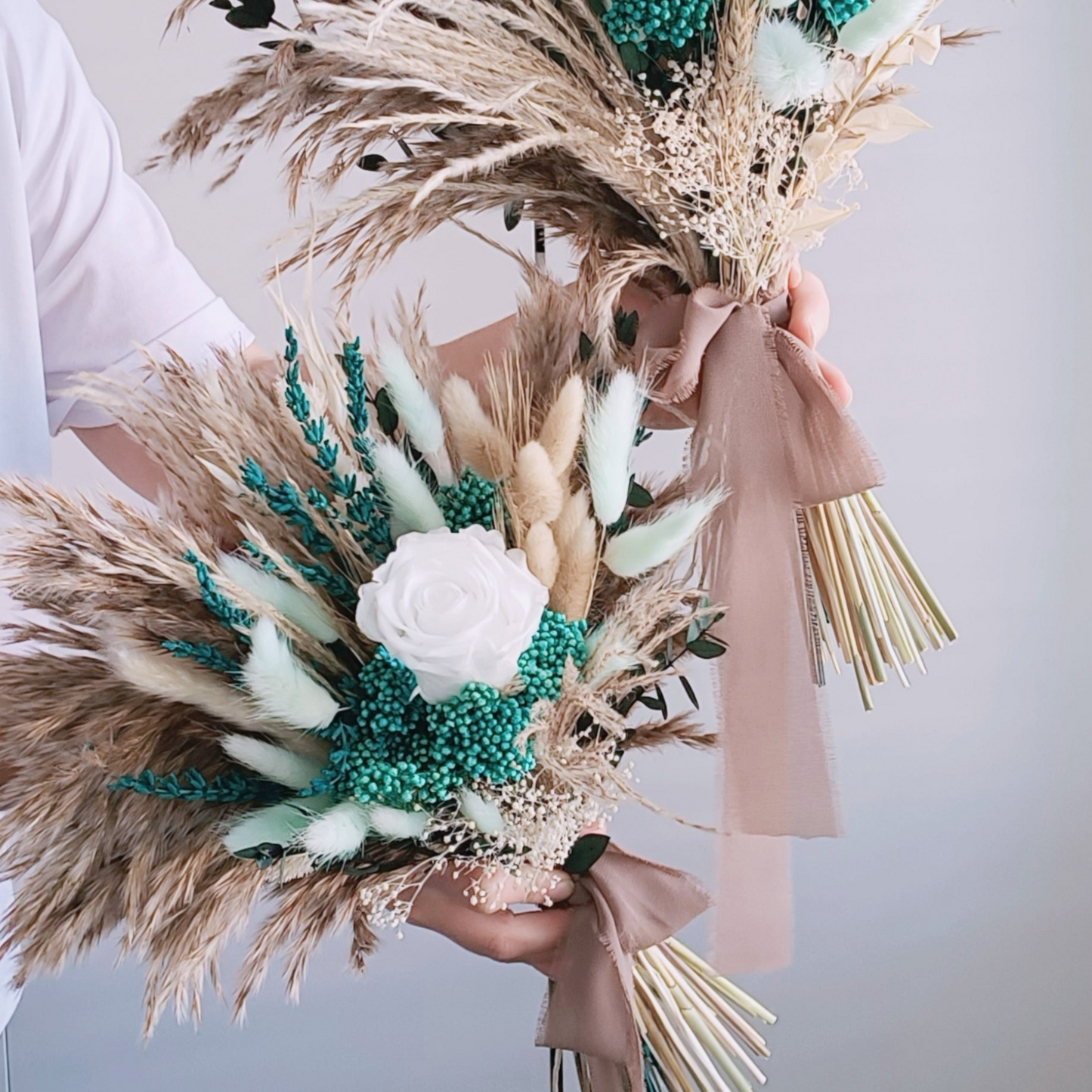 Featuring a sophisticated mix of teal and white, this dried flower bouquet is a striking choice for the modern boho bride. Pampas grass and preserved foliage add a touch of natural elegance, while the teal accents bring a fresh and vibrant feel. Perfect for rustic-themed weddings, this long-lasting bouquet is designed to make a lasting impression. Discover more teal and white bridal bouquets in our exclusive collection.