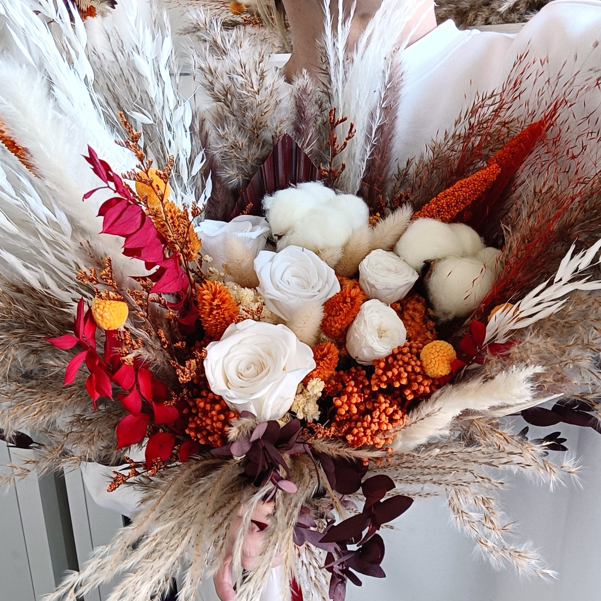 This eye-catching wedding bouquet features a bold combination of red, orange, and natural hues, crafted from a selection of dried flowers. The arrangement includes pampas grass, preserved foliage, and textured accents, offering a rustic yet vibrant look perfect for autumn weddings. Long-lasting and full of character, this bouquet adds a fiery touch to any bridal ensemble. Explore our collection for more unique and seasonal wedding bouquets.