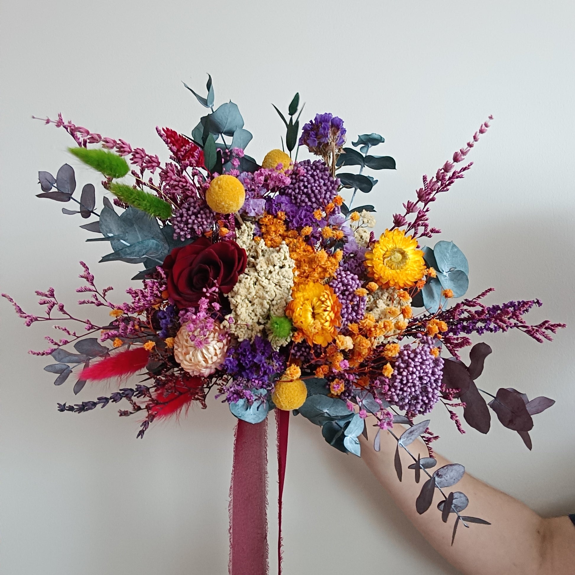 Vibrant dried flower bridal bouquet in jewel tones, featuring preserved burgundy sola rose, yellow billy buttons, purple statice, orange strawflowers, red celosia, and eucalyptus foliage in blue and plum shades. Handheld against a plain wall, with a long deep red ribbon. Bold, colourful design perfect for boho, rustic, or artistic weddings. Rich texture and colour make it ideal for standout bridal styling.