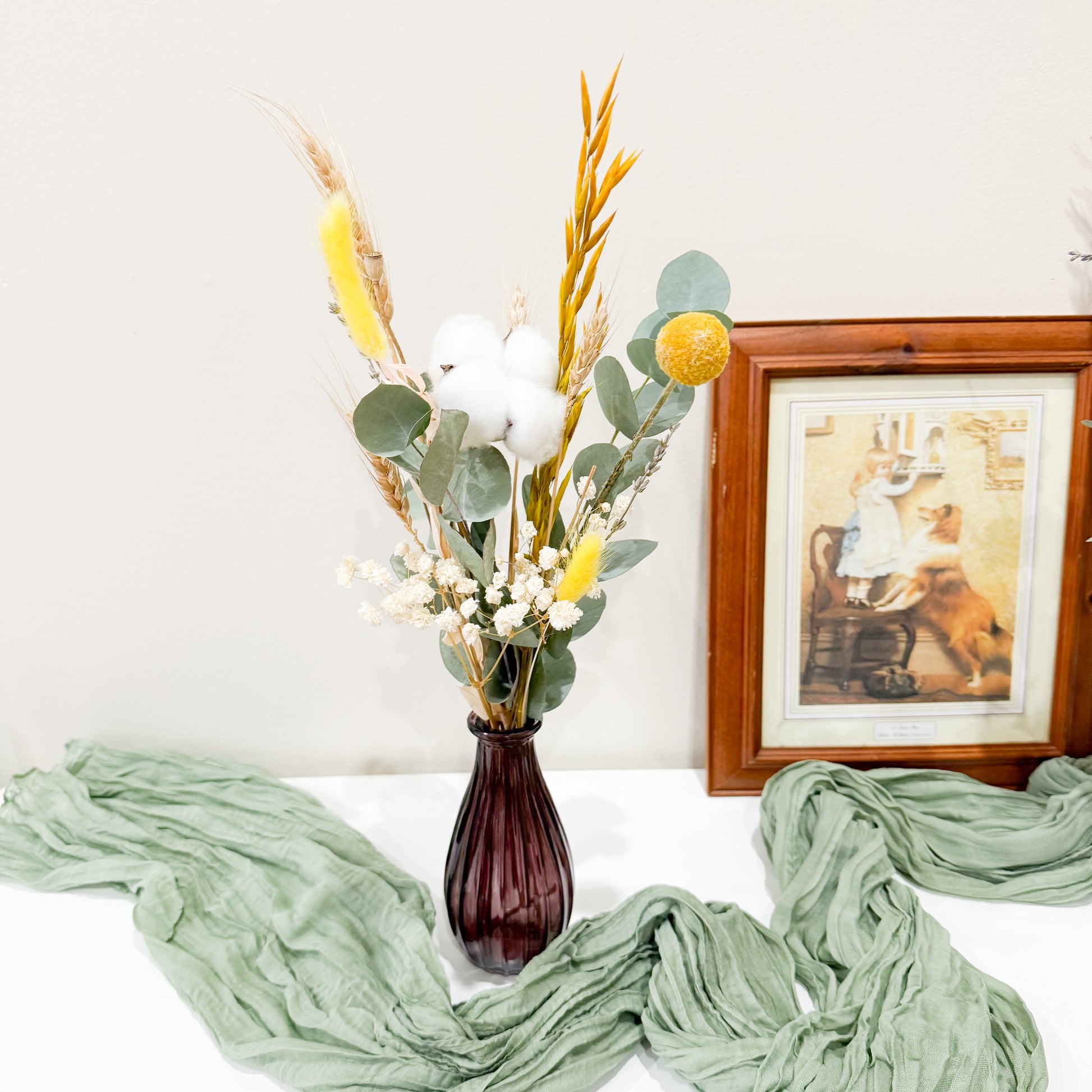 Boho dried flower arrangement in brown vase with preserved eucalyptus, white cotton, yellow bunny tails, and natural textures on a soft green drape.