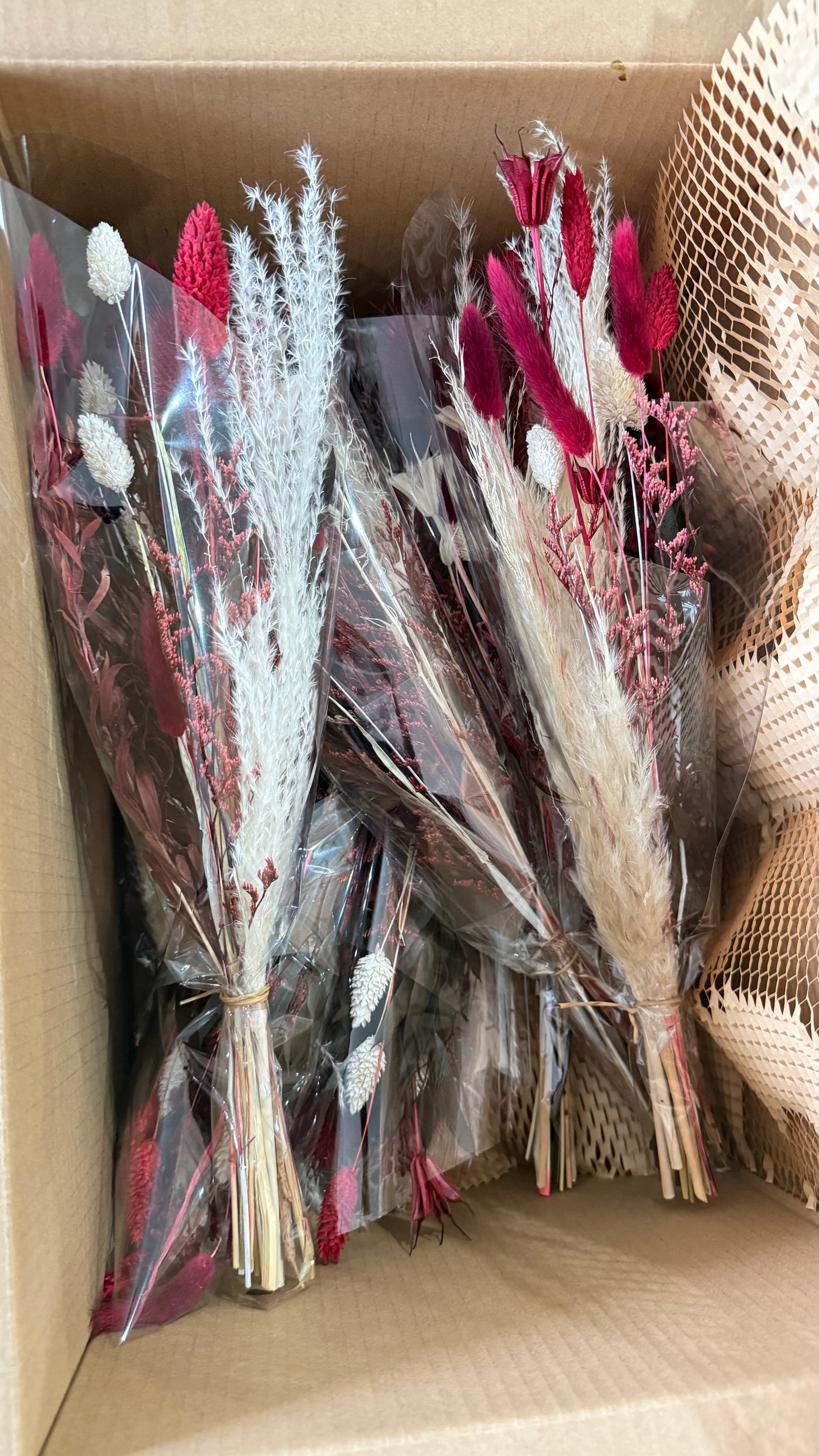 Several small dried flower bouquets wrapped in clear plastic inside a cardboard box. The bouquets feature white, pink, and burgundy dried flowers with beige grasses.