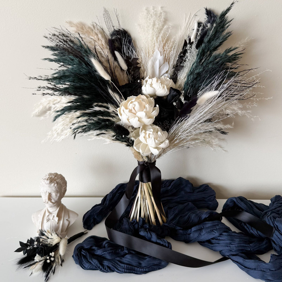 A dried floral arrangement featuring pampas grass in black, white, and dark green tones, with cream-colored flowers and a black ribbon, displayed on a white surface next to a small white bust and draped navy fabric.
