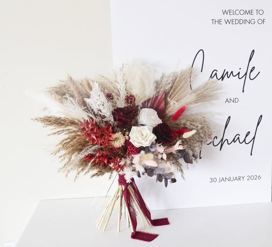 Dried wedding bouquet in burgundy and ivory tones placed in front of a welcome wedding sign – pampas, roses, and preserved florals.