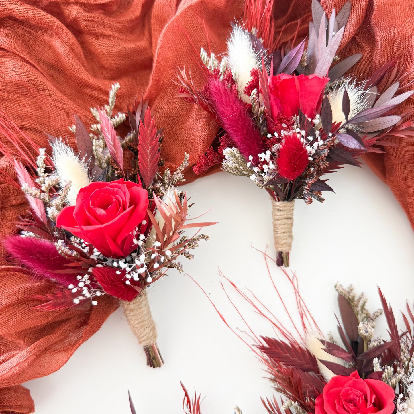Boho wedding dried flower boutonnieres in red and burgundy tones, sola wood roses with rustic wrap, handmade groomsmen accessories for rustic and bohemian weddings.