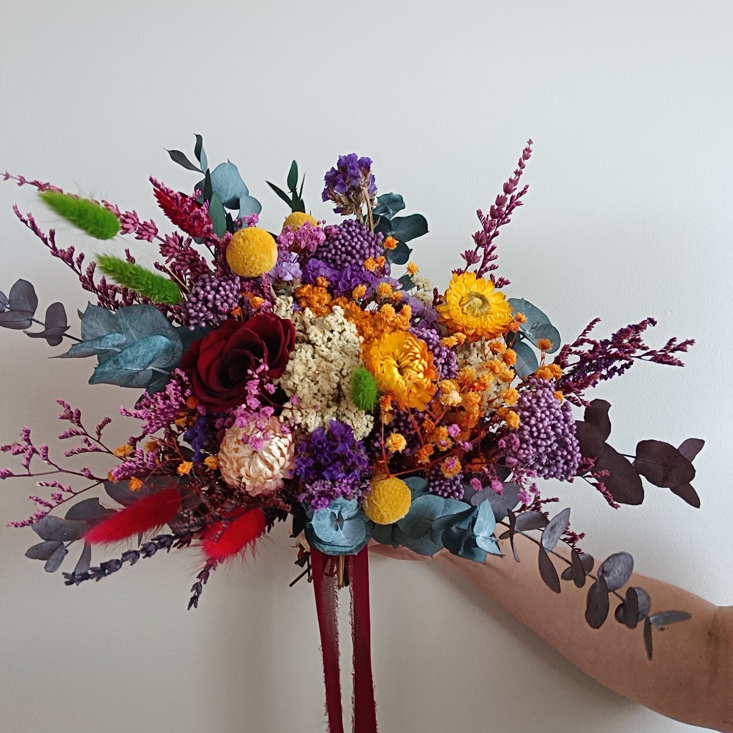 Side view of a colourful dried flower bouquet in jewel tones. Includes preserved blooms in deep burgundy, golden yellow, lilac, blue-green, and crimson, arranged with eucalyptus and accent foliage. Tied with a dark red ribbon and held against a white background. Perfect for brides seeking a unique, vibrant wedding bouquet with bold colours and a wild, natural shape.
