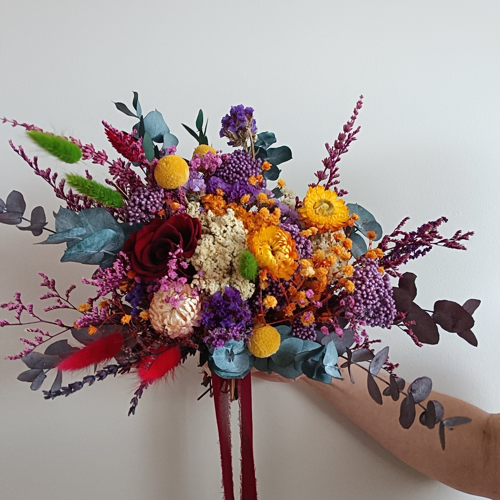 Side view of a colourful dried flower bouquet in jewel tones. Includes preserved blooms in deep burgundy, golden yellow, lilac, blue-green, and crimson, arranged with eucalyptus and accent foliage. Tied with a dark red ribbon and held against a white background. Perfect for brides seeking a unique, vibrant wedding bouquet with bold colours and a wild, natural shape.