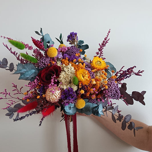 Side view of a colourful dried flower bouquet in jewel tones. Includes preserved blooms in deep burgundy, golden yellow, lilac, blue-green, and crimson, arranged with eucalyptus and accent foliage. Tied with a dark red ribbon and held against a white background. Perfect for brides seeking a unique, vibrant wedding bouquet with bold colours and a wild, natural shape.