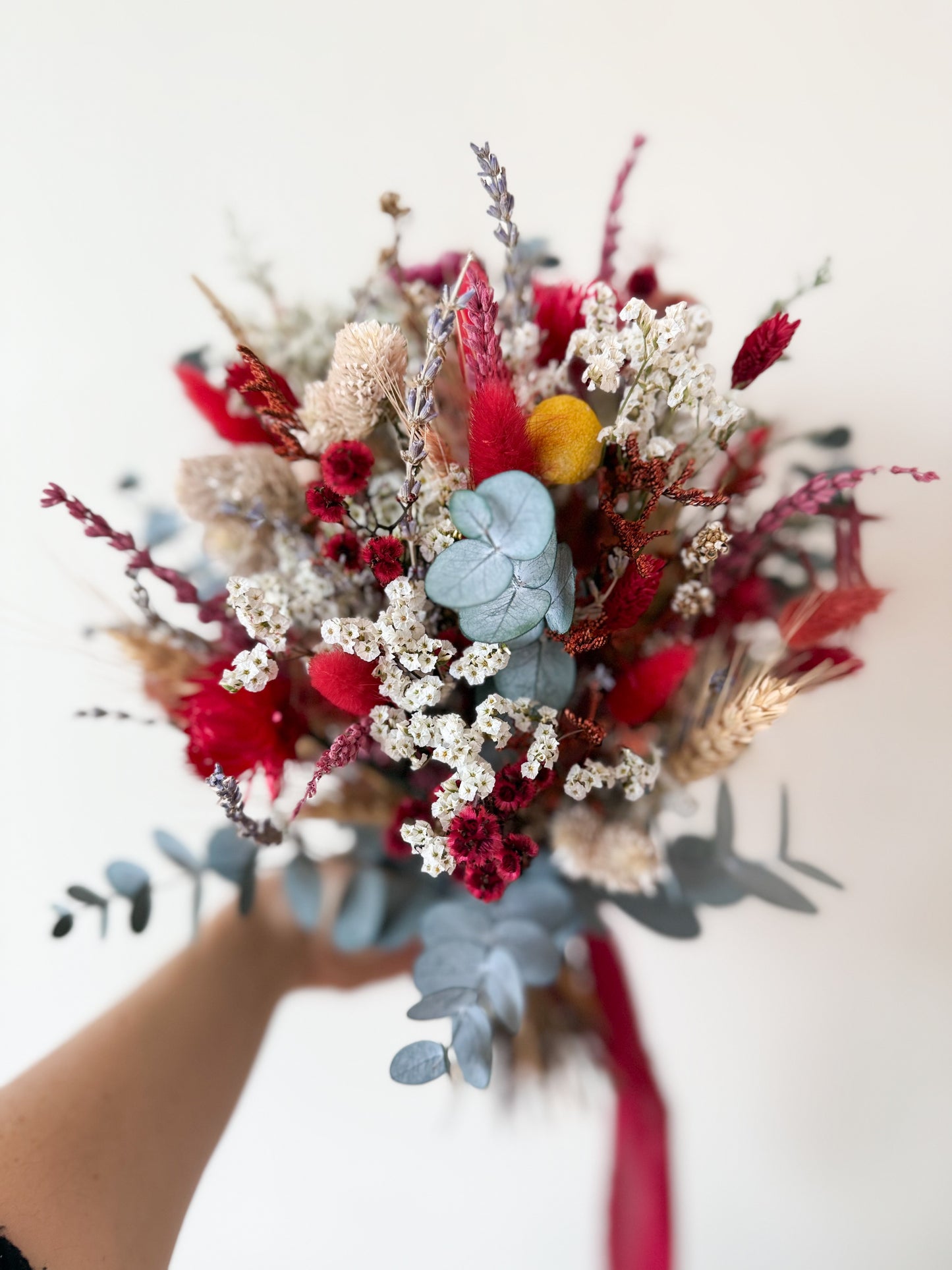 Dried boho wedding bouquet in red, white and blue eucalyptus held against a cream background
