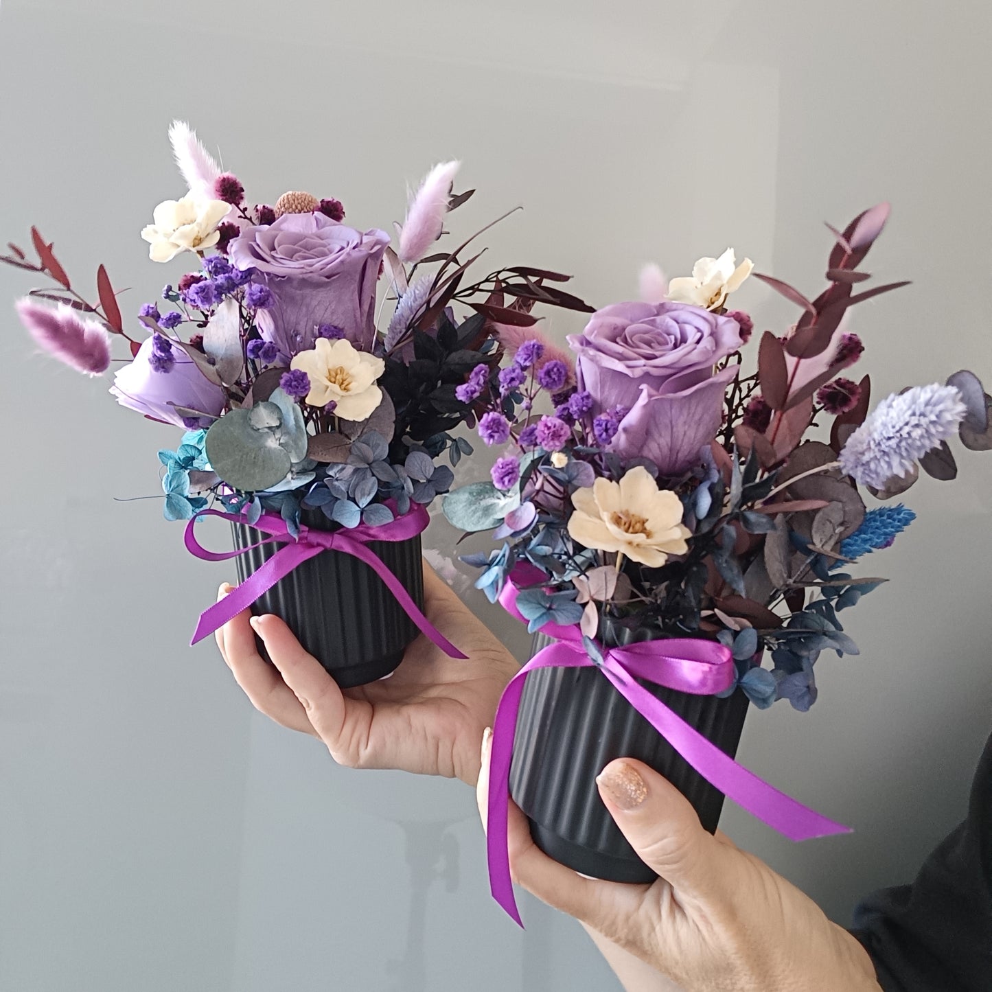 Two small floral arrangements with purple and white flowers held by hands against a gray background