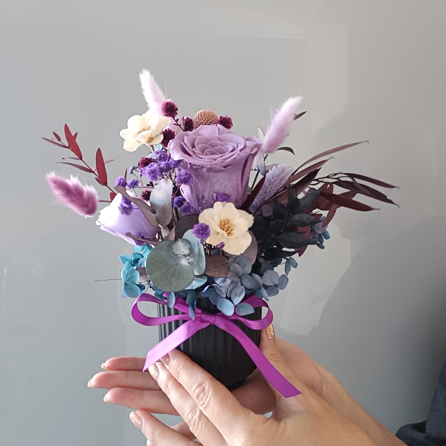 Hand holding a small bouquet of flowers with a purple ribbon against a plain background