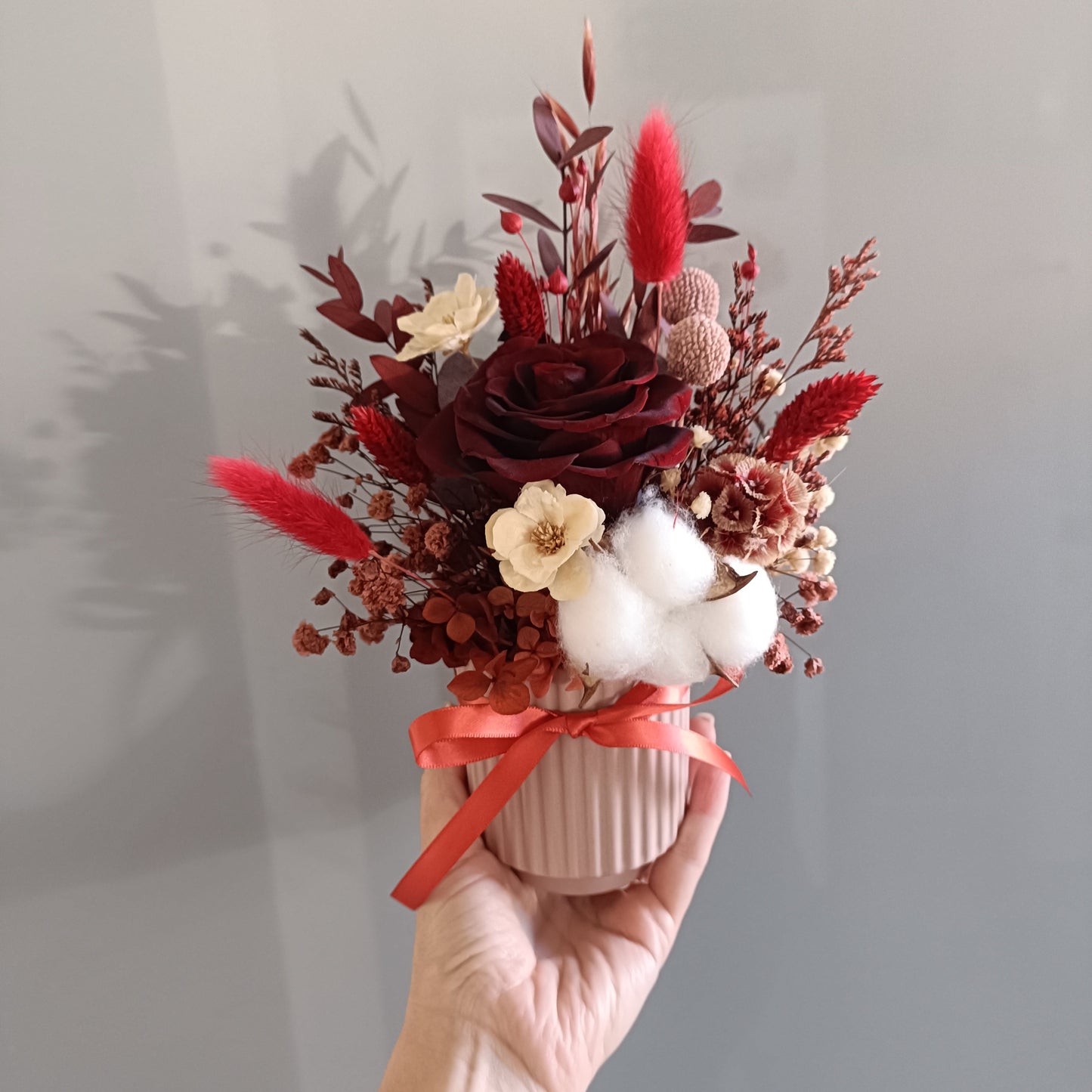 Single dried flower arrangement held in hand. Features a deep red preserved rose, white cotton, sola flowers, dried red bunny tails, and natural preserved filler. Tied with a red satin ribbon and placed in a ceramic ribbed vase. A romantic and sustainable floral gift idea with a warm, passionate colour palette.