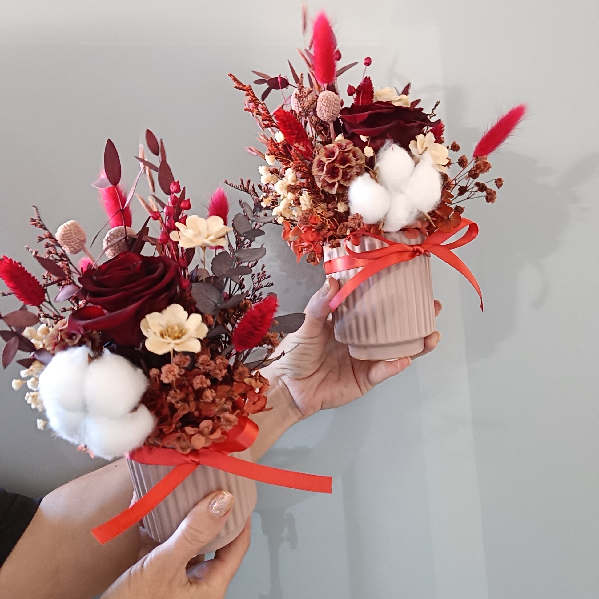 Two dried flower arrangements in red, cream, and burgundy tones displayed side by side. Each bouquet features a preserved red rose, sola flowers, white cotton, red bunny tails, and rustic filler in a ribbed pink ceramic vase, tied with a red ribbon. Ideal for romantic gifts, Valentine’s Day, or stylish home decor. Elegant, long-lasting floral pieces made with preserved and dried botanicals.