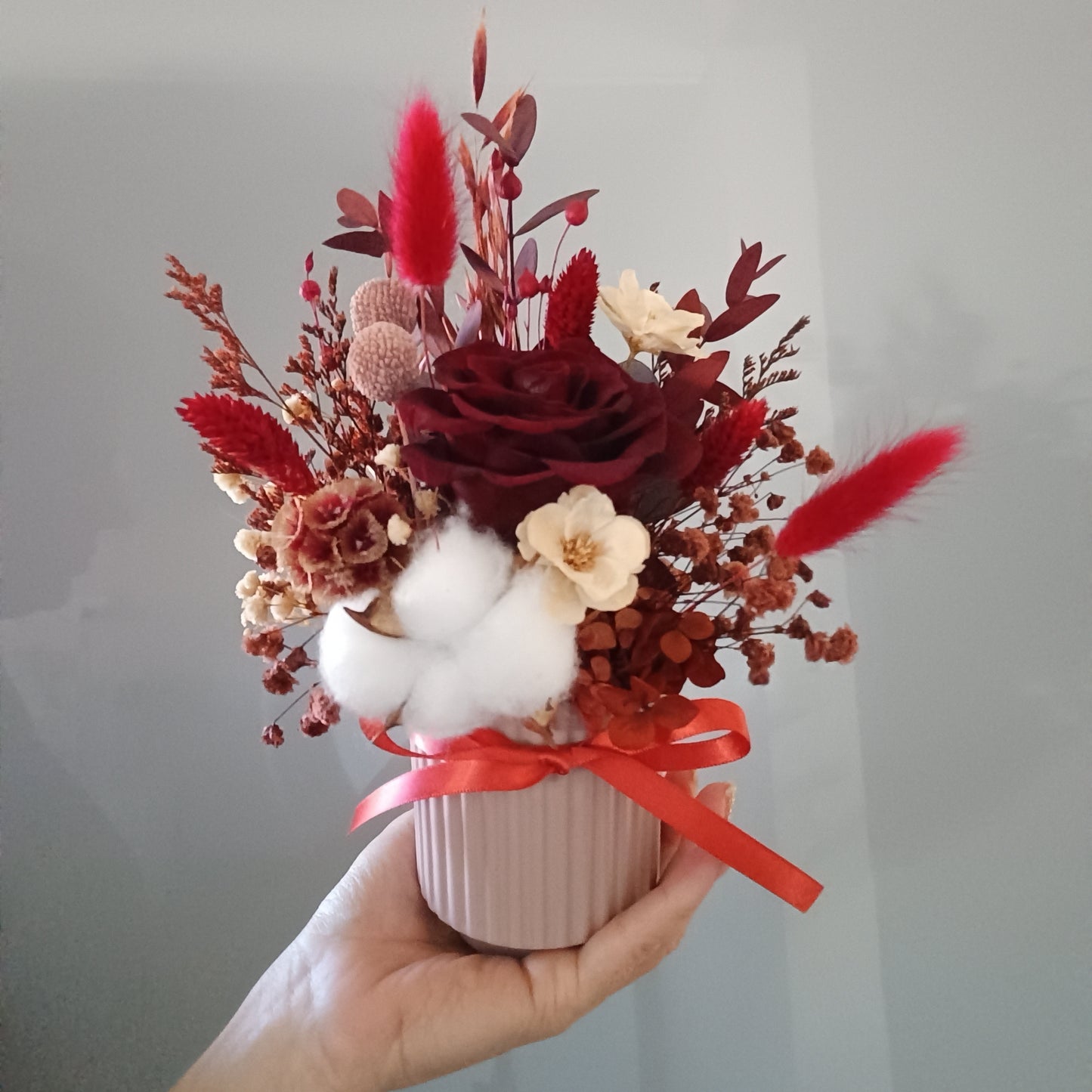 Close-up view of a mini dried bouquet in rich red and neutral tones. Includes preserved rose, sola wood flowers, cotton pods, red foliage, and dried berries. Perfect as a desk arrangement, thank-you gift, or Valentine’s decoration. The soft textures and vibrant colours offer lasting beauty without maintenance