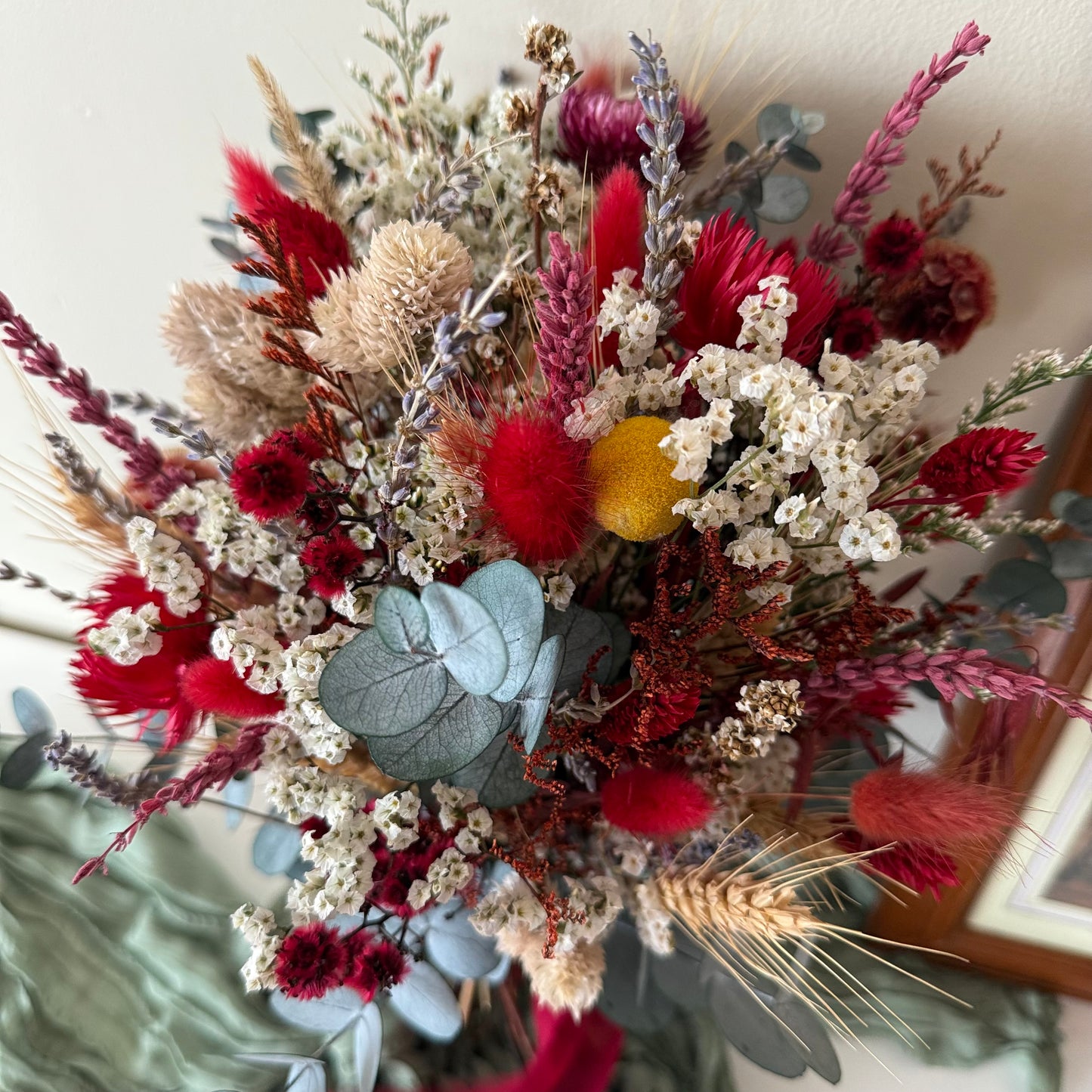Red and white dried wedding bouquet on sage-green fabric with framed artwork in background