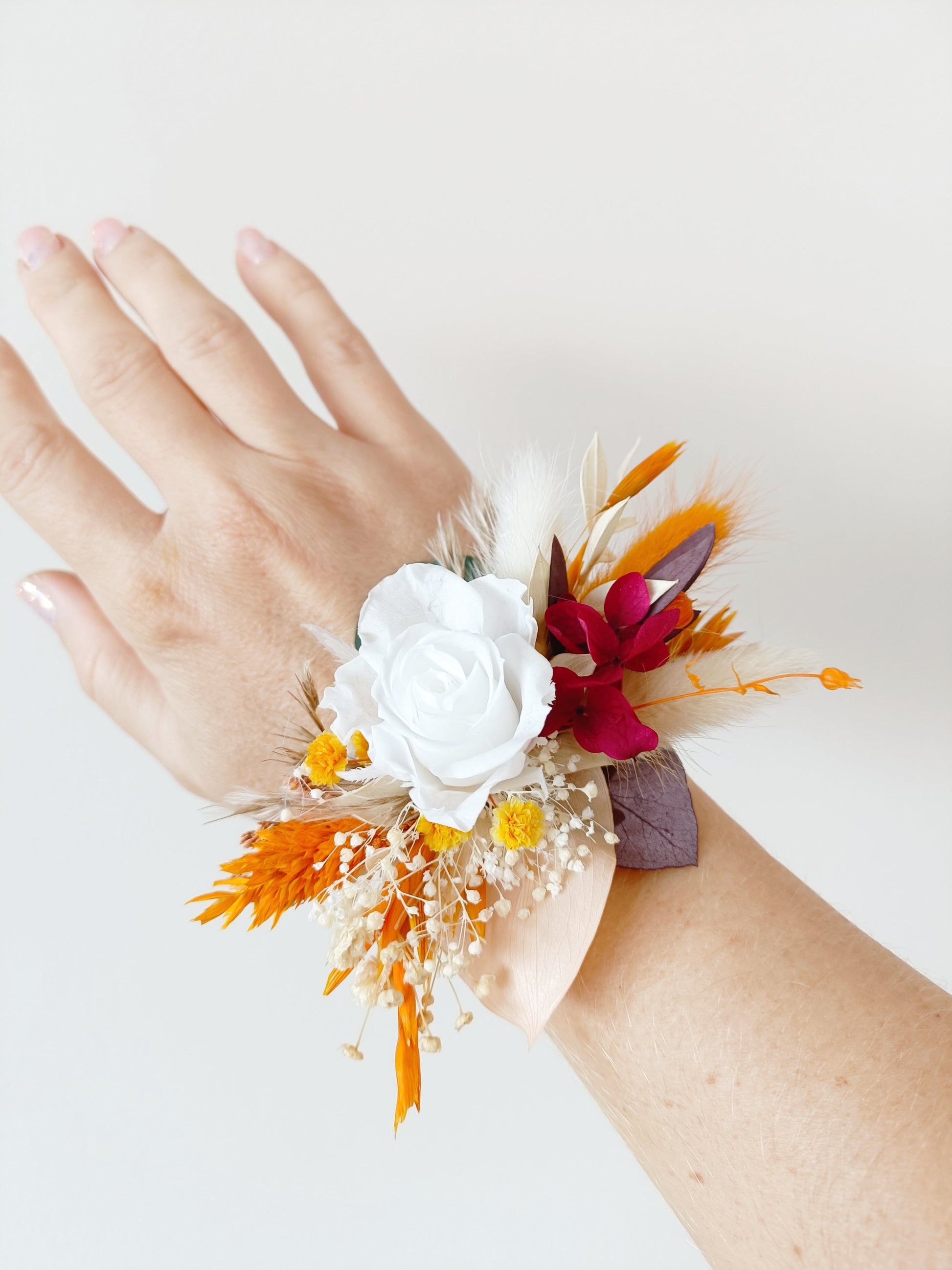 Wrist corsage with white preserved rose, orange accents, and burgundy dried flowers, worn on a hand.