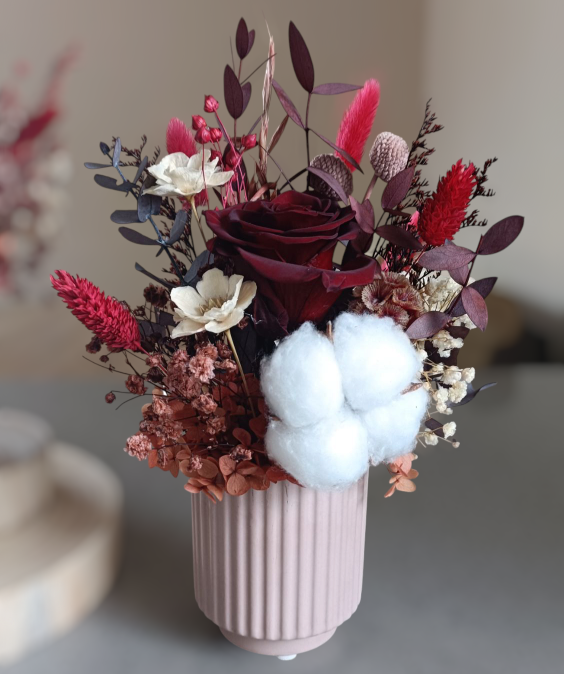 Overhead view of preserved floral arrangement in a white ceramic vase. Floral mix includes a deep red rose, cream sola flowers, cotton, bunny tails, and preserved leaves in warm red and burgundy tones. A compact, romantic floral piece for gifts, bedside tables, or modern interiors.