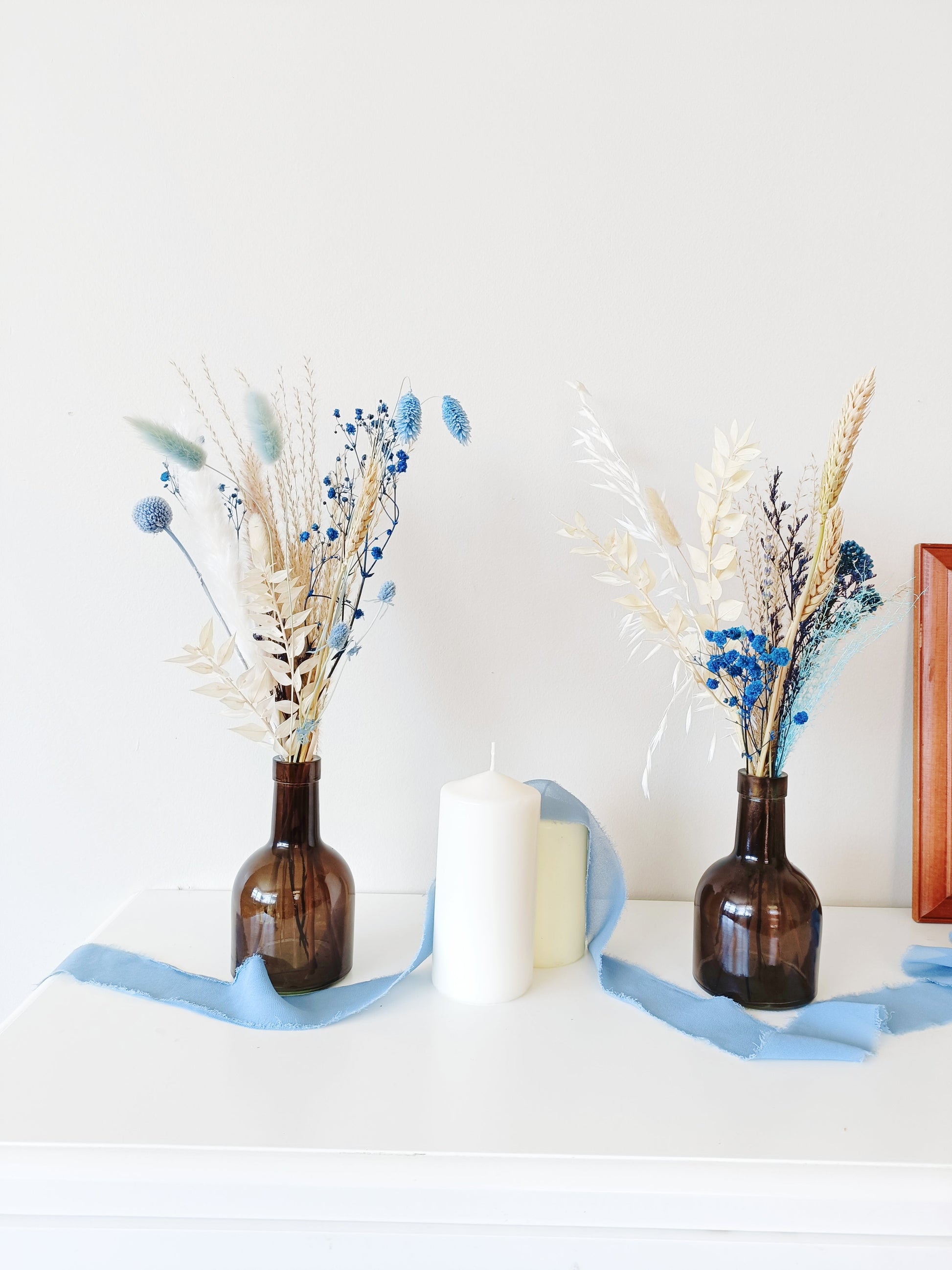 Pair of dried flower arrangements in amber glass vases with blue and beige preserved blooms – styled with candles and dusty blue ribbon for modern rustic decor.