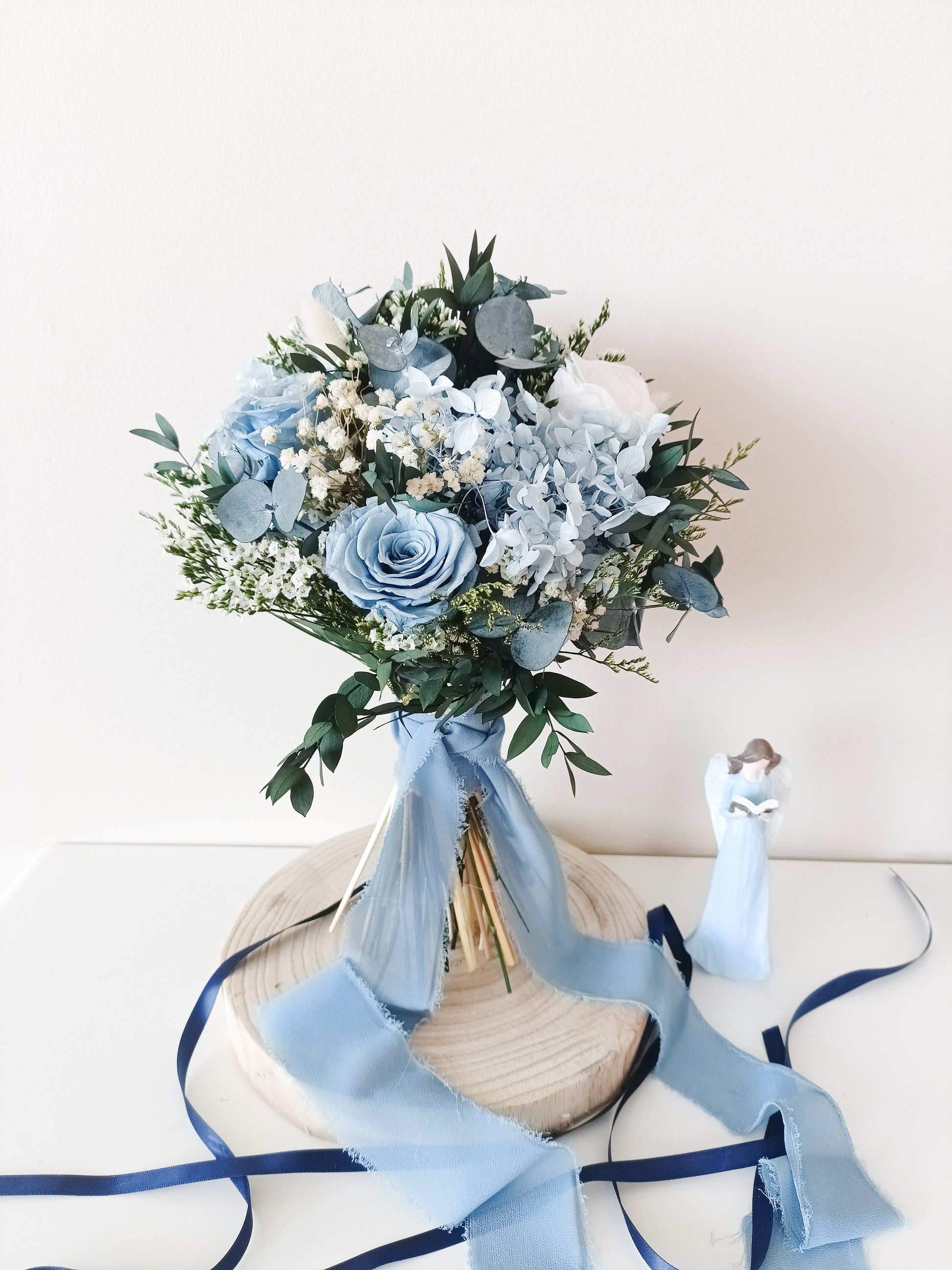 Hand-tied bouquet in dusty blue shades with preserved flowers and trailing velvet ribbon, arranged on a wooden stand next to a white angel figurine.