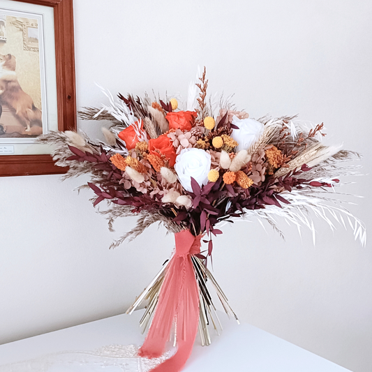 Elegant terracotta-inspired dried flower wedding bouquet with warm orange roses, fluffy pampas grass, and preserved burgundy greenery – styled on a clean white background with dusty pink ribbon.