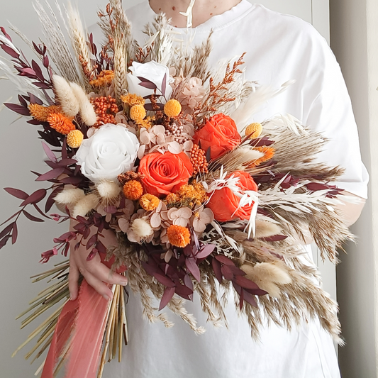 Bold dried flower bridal bouquet in burnt orange, burgundy, and ivory tones featuring sola wood roses, pampas grass, craspedia, and preserved foliage – held by bride in white top, styled for rustic or boho weddings.