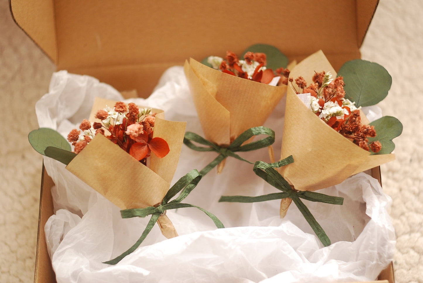 A close-up of a single dried flower bouquet in a box, wrapped in kraft paper with green ribbon and accompanied by eucalyptus leaves. Flowers in shades of terracotta, burnt orange, and white. The soft fabric lining enhances the gift presentation. A thoughtful choice for bridesmaid gifts or sustainable floral favours.