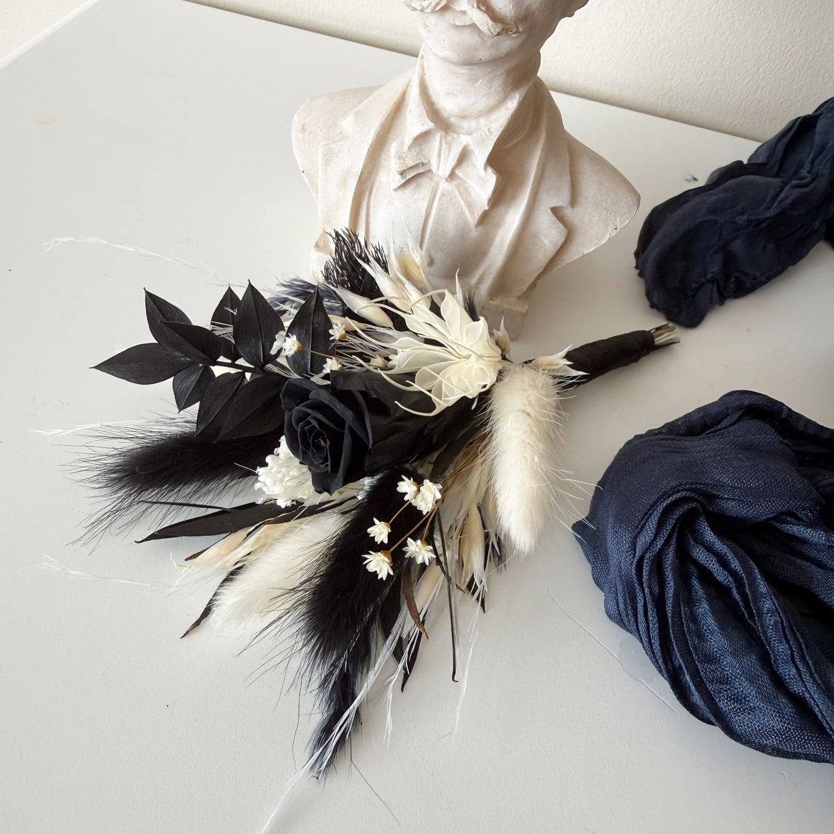 A gothic-style bouquet featuring black and cream feathers, dried white flowers, and black leaves, styled in front of a white bust sculpture with navy blue fabric rolls on a white surface.