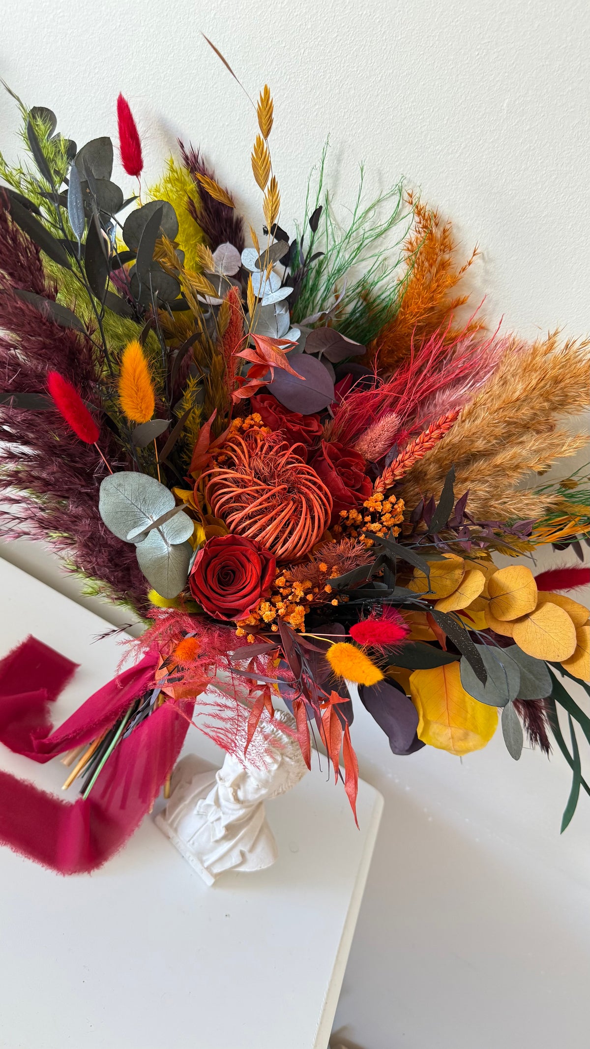 A dried floral arrangement featuring autumnal elements including red roses, eucalyptus, and orange foliage, tied with a magenta ribbon and displa
