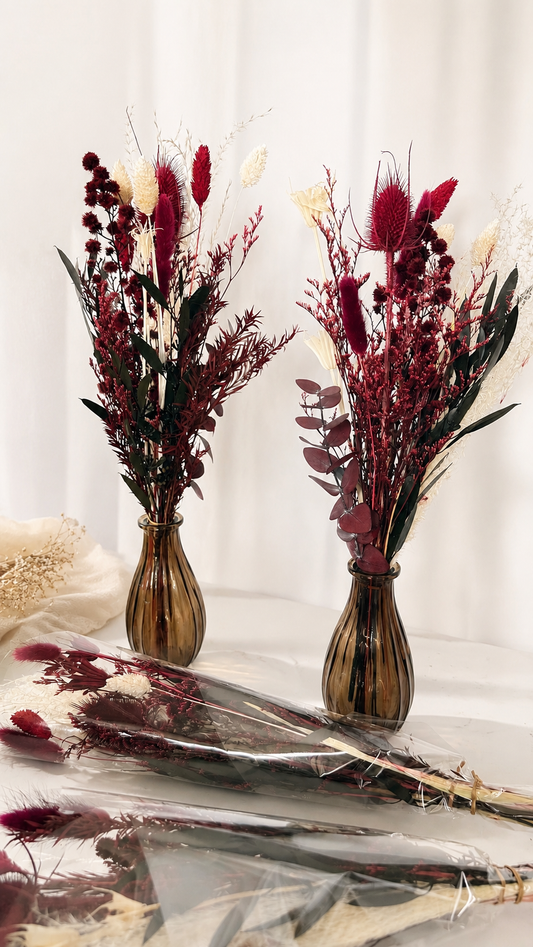 Two amber glass vases filled with dried floral arrangements in deep burgundy, burgundy-red, cream, and dark green tones, displayed on a draped white fabric surface.