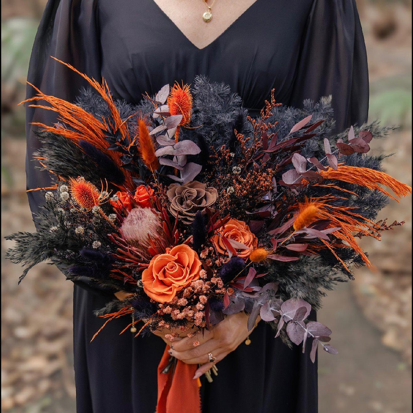 Bridal bouquet in burnt orange and black tones featuring preserved roses, Australian native flowers, textured foliage, and dried grasses, held by a woman in a black dress for a modern boho wedding.
