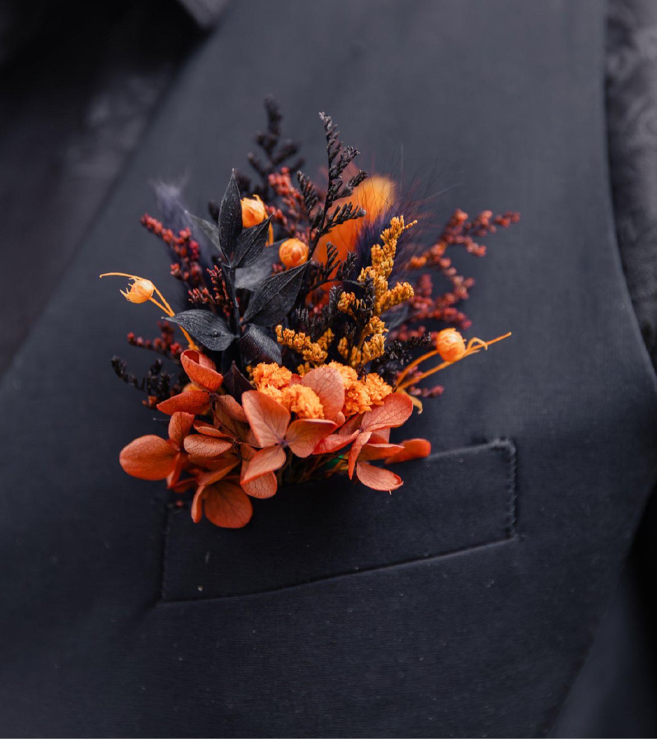 Close-up of a burnt orange and black dried flower boutonniere with preserved hydrangea, Australian native accents, and textured foliage, styled on a black suit jacket for a modern boho wedding.