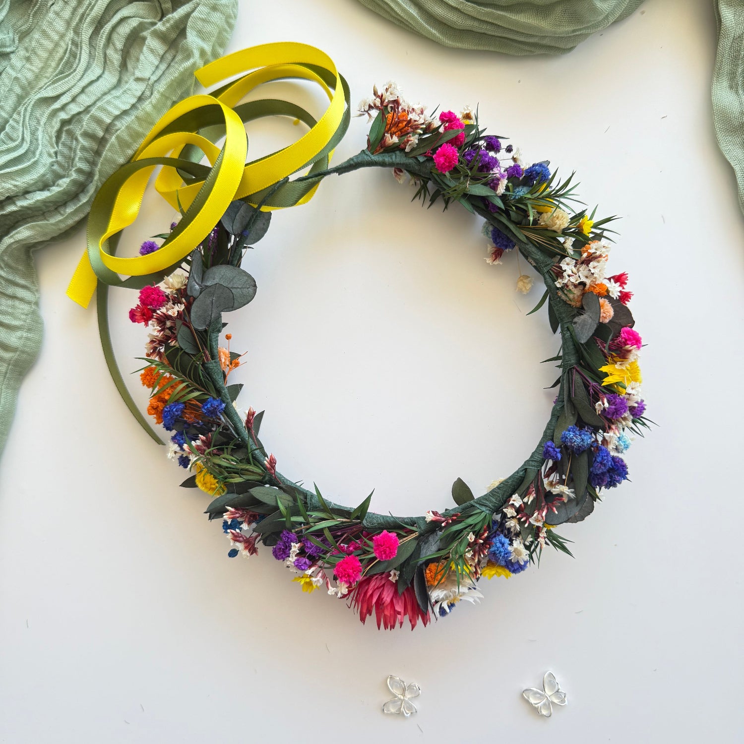Handmade dried floral crown featuring red strawflower, blue statice, white daisies and eucalyptus leaves tied with yellow ribbon.