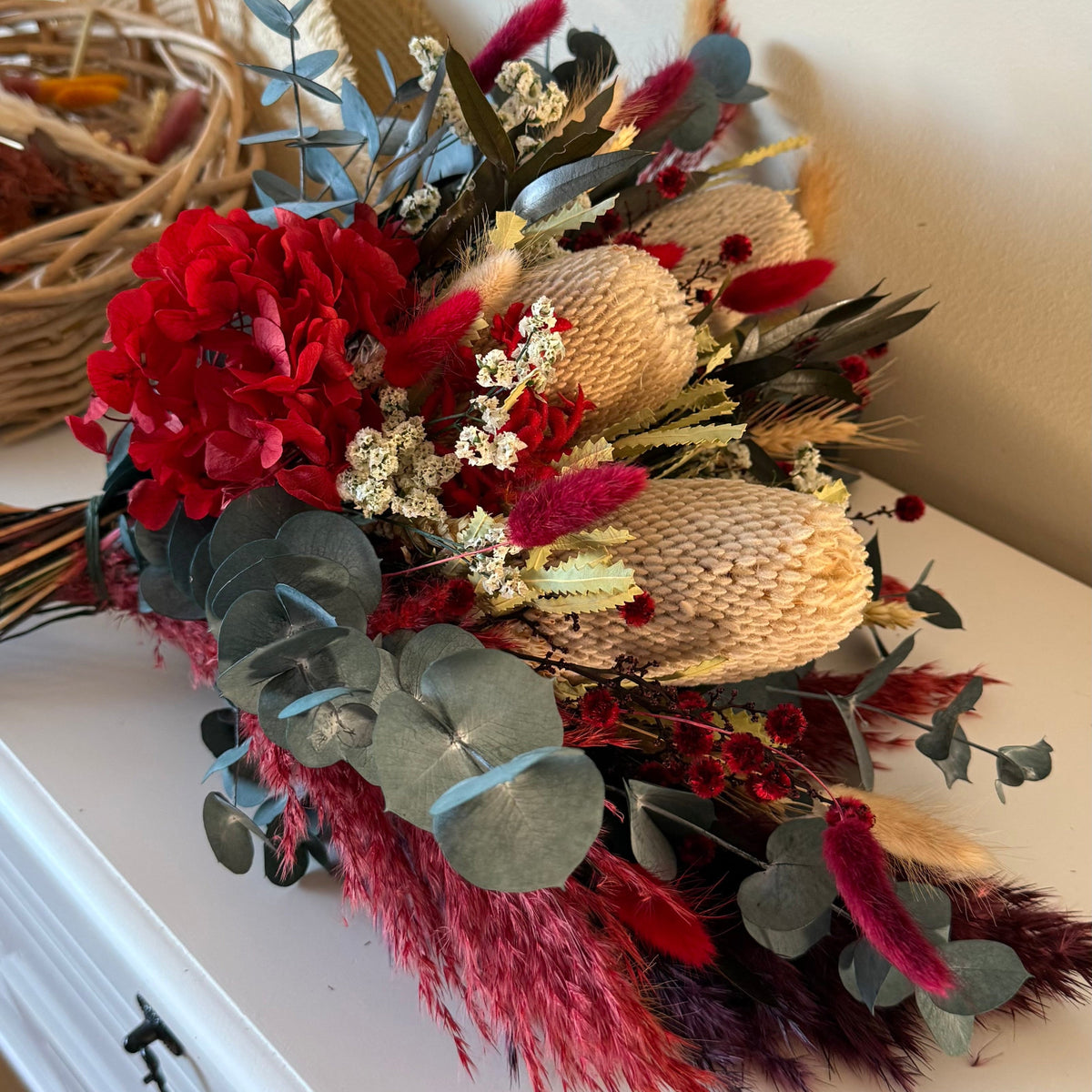 A dried floral arrangement featuring vibrant red flowers, beige protea blooms, green eucalyptus leaves, and red pampas grass, styled on a white wooden cabinet.