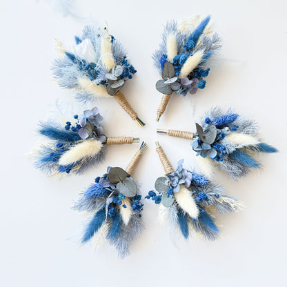 Round flat lay of dried boutonniere set with preserved bunny tails, blue blooms and ivory accents for groomsmen or bridesmaids.