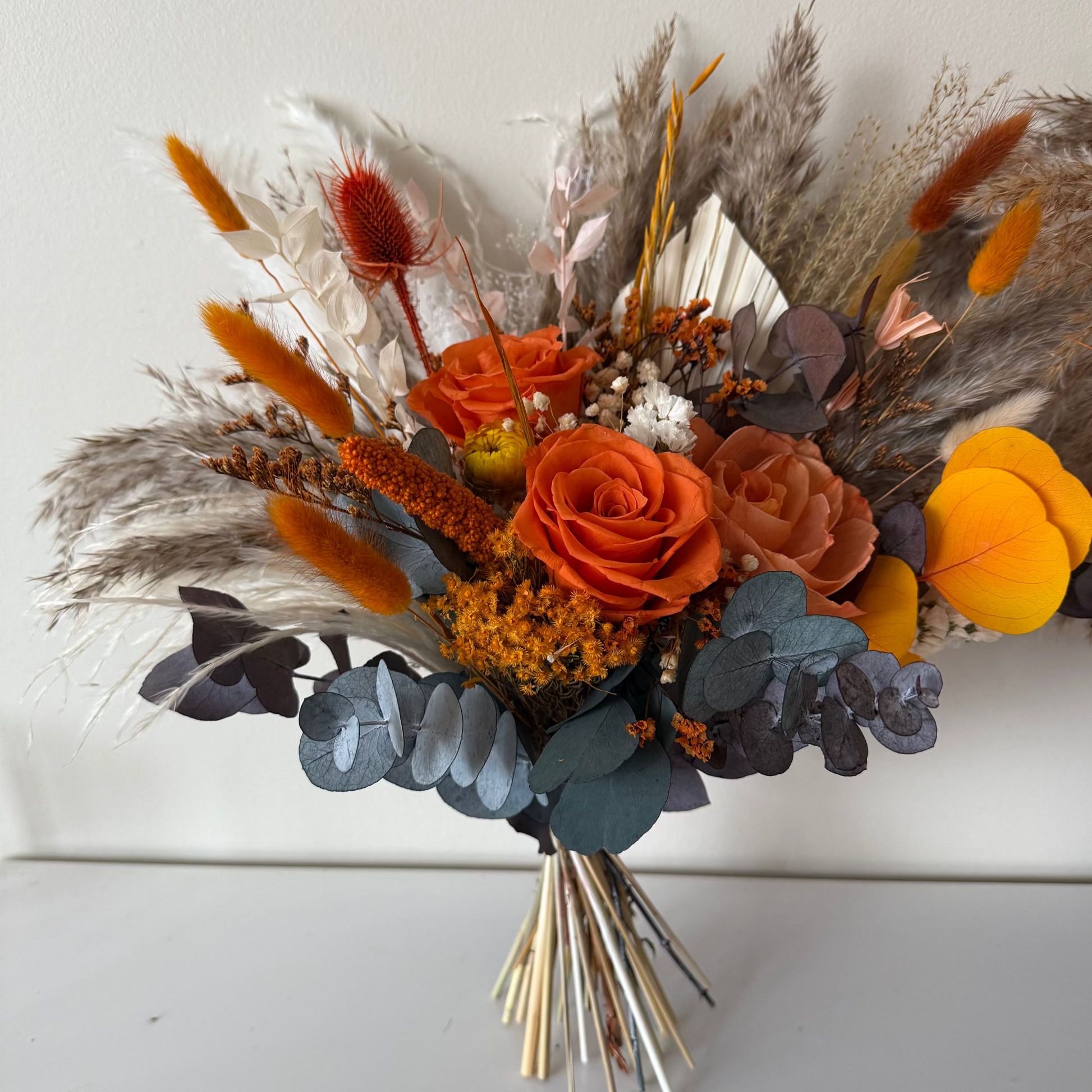 Flat lay image of a boho bridal bouquet with terracotta, orange, and yellow dried flowers, styled with green fabric and a peach candle for a warm wedding aesthetic.
