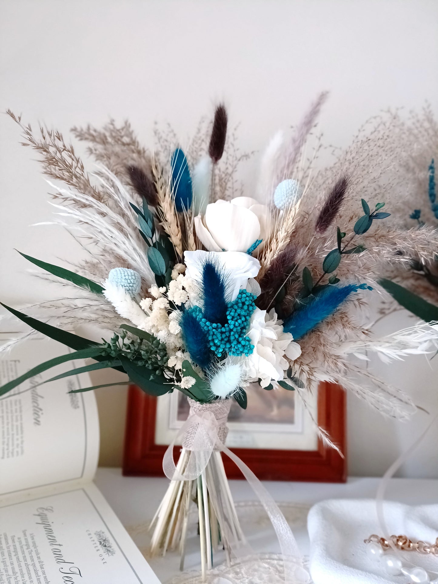 This handcrafted wedding bouquet features an exquisite blend of dried flowers, combining soft natural tones with striking blue accents. The arrangement includes pampas grass, preserved foliage, and delicate bunny tails, creating a perfect balance of rustic charm and modern elegance. Ideal for a boho-themed wedding, this long-lasting bouquet adds a touch of timeless beauty to your special day. Explore our collection for more unique bridal bouquets and wedding decor ideas.